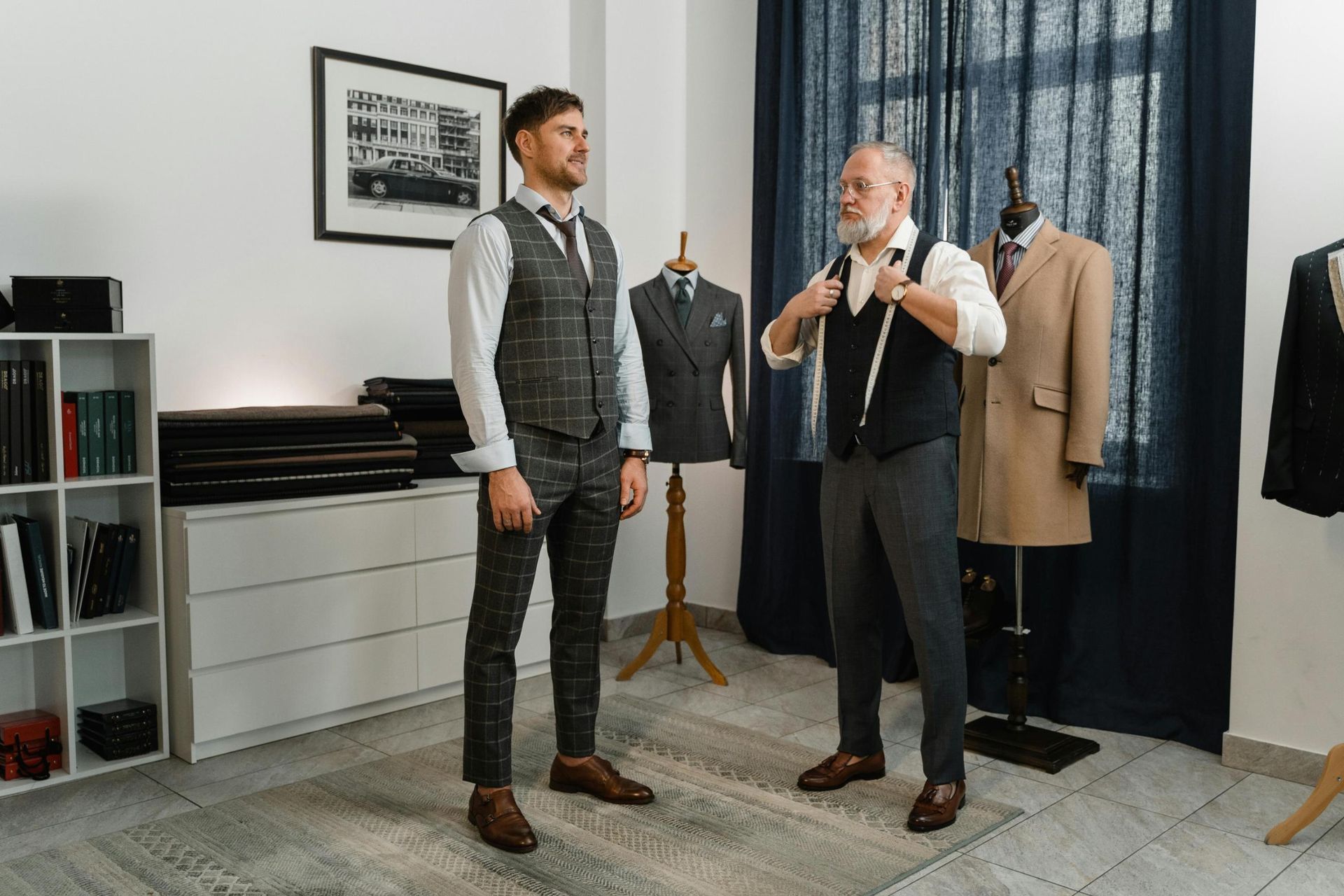 In a professional tailoring shop, an older tailor with a measuring tape around his neck stands opposite a client wearing a three-piece windowpane suit. The studio features organized fabric bolts, a framed car photograph, and several mannequins displaying well-crafted menswear.