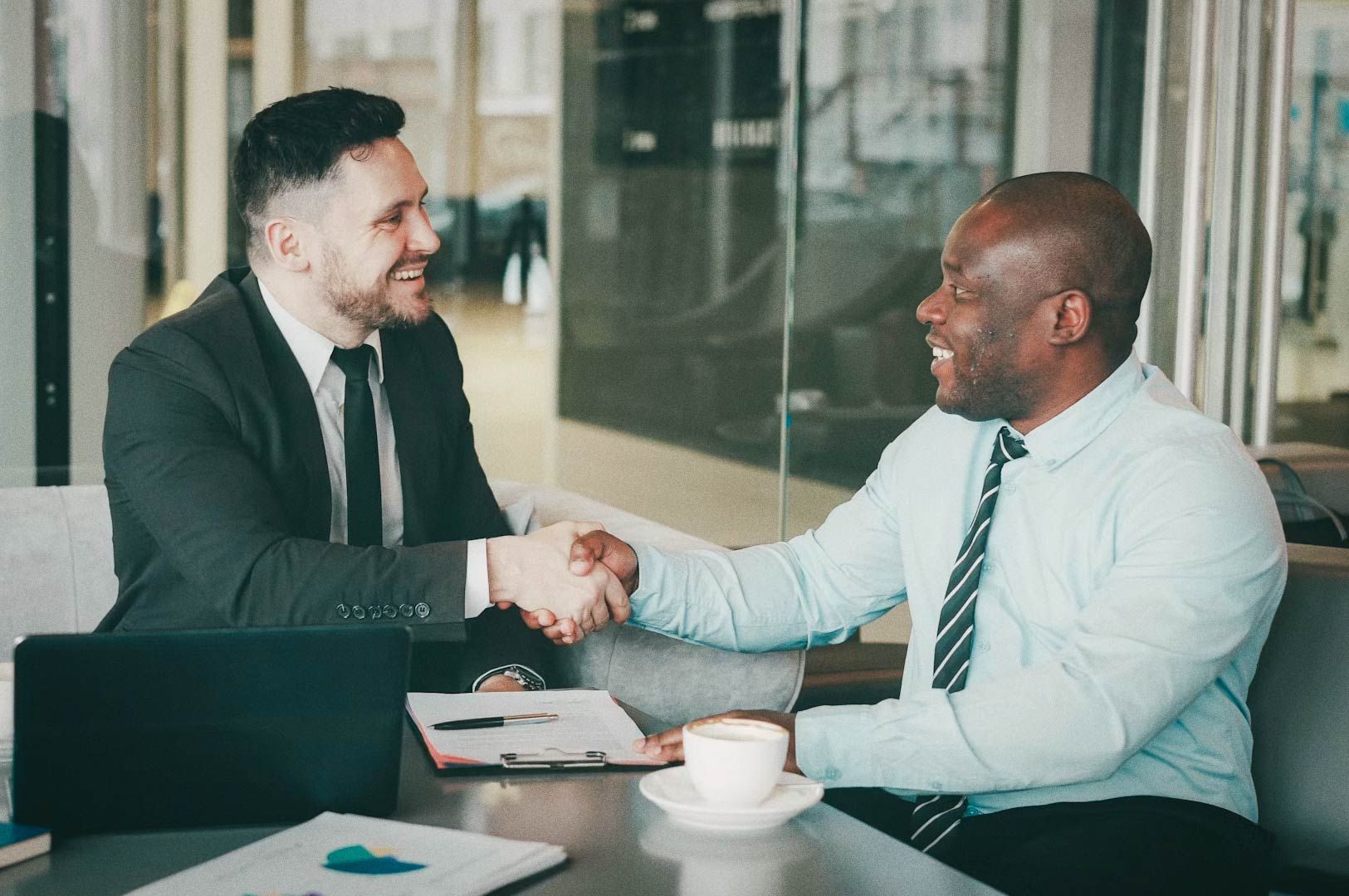 Two professionals in suits shaking hands across table with laptop and documents in modern office setting.