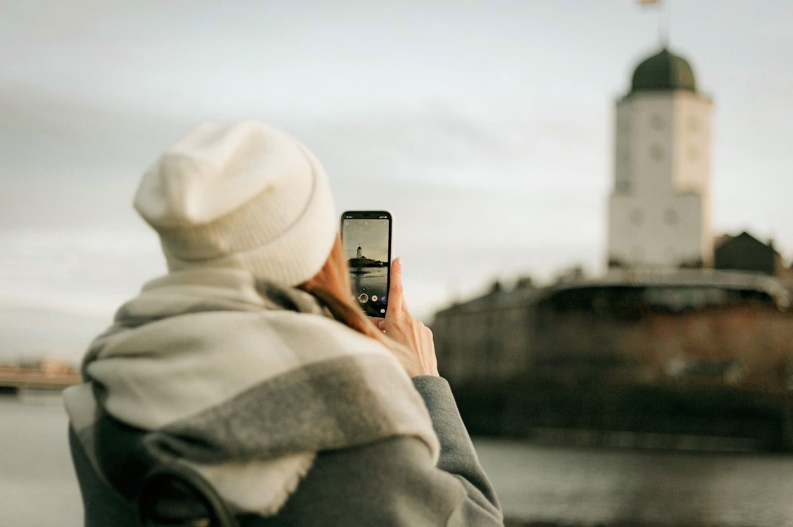 A traveler taking a photo of a historic waterfront tower with a smartphone during a cold, overcast day.