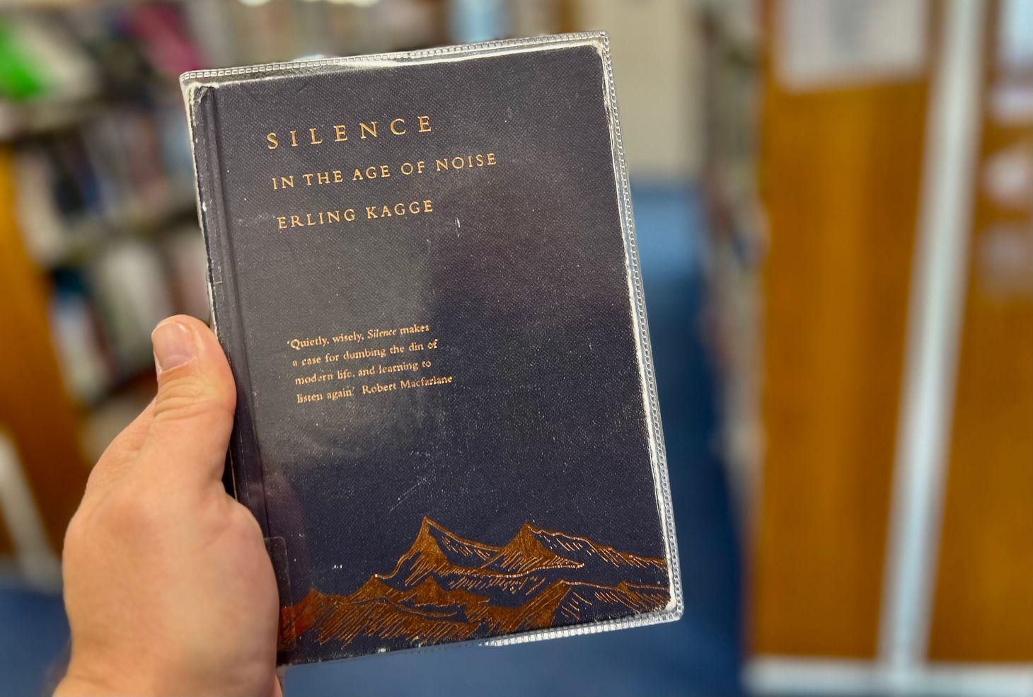 A person's hand holds a small, navy blue book titled Silence: In the Age of Noise by Erling Kagge, w