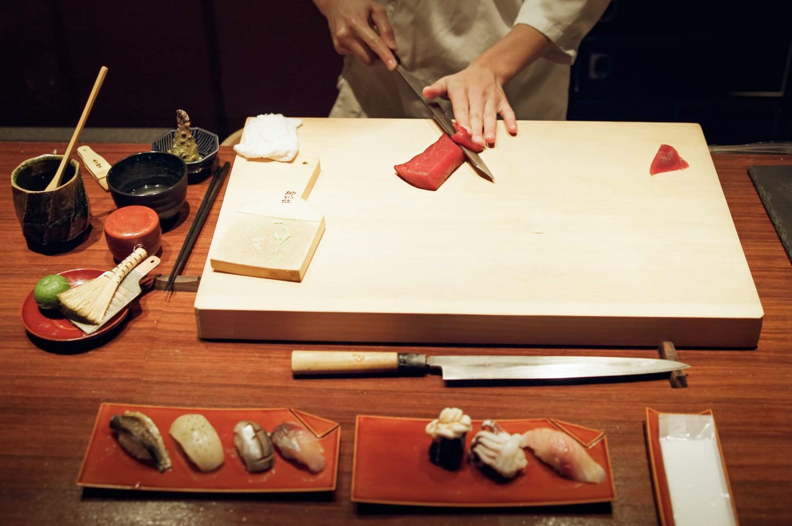 Chef slicing a piece of fresh tuna on a wooden sushi counter with sushi knives, small bowls, and assorted nigiri arranged in front.
