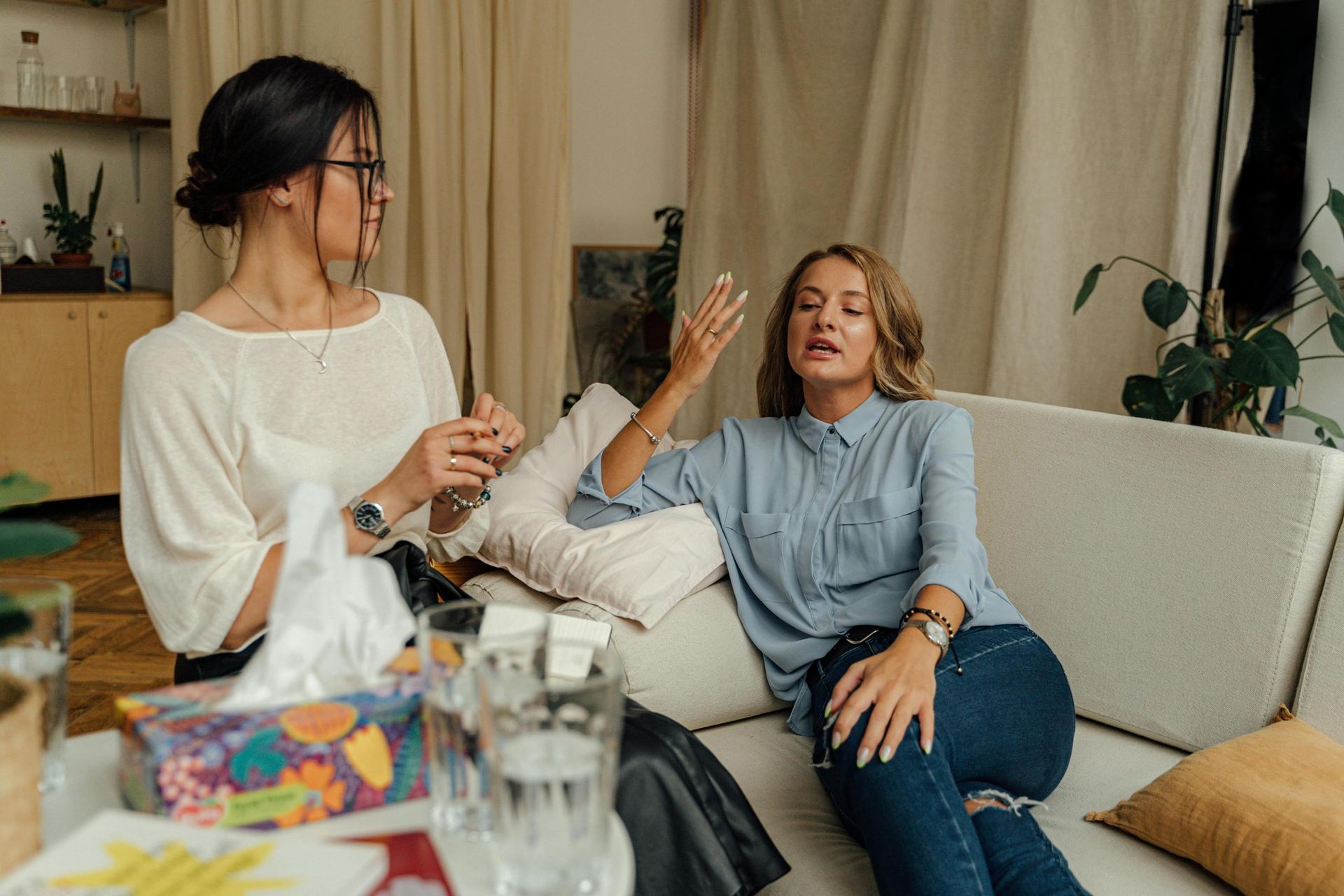 A woman in a blue shirt speaks expressively with hand gestures while sitting on a sofa next to an attentive listener wearing glasses. In the foreground, a box of tissues and glasses of water rest on a table, indicating a therapy or counseling setting.