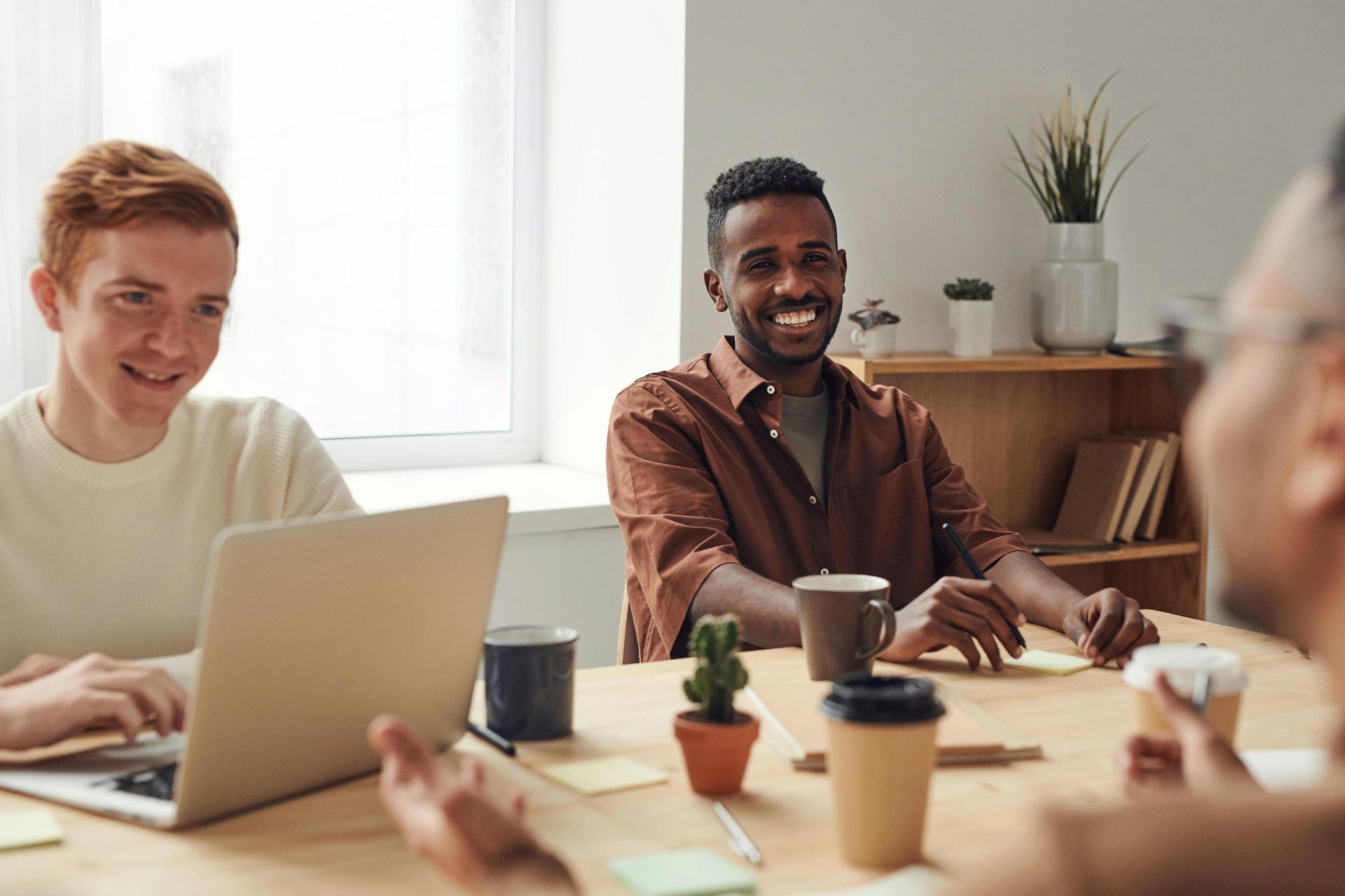 Three men are gathered at a wooden table with laptops, sticky notes, and coffee cups in a bright office environment. The central figure smiles warmly at a colleague across from him, while a man on the left types on his computer, indicating a collaborative and positive work session.