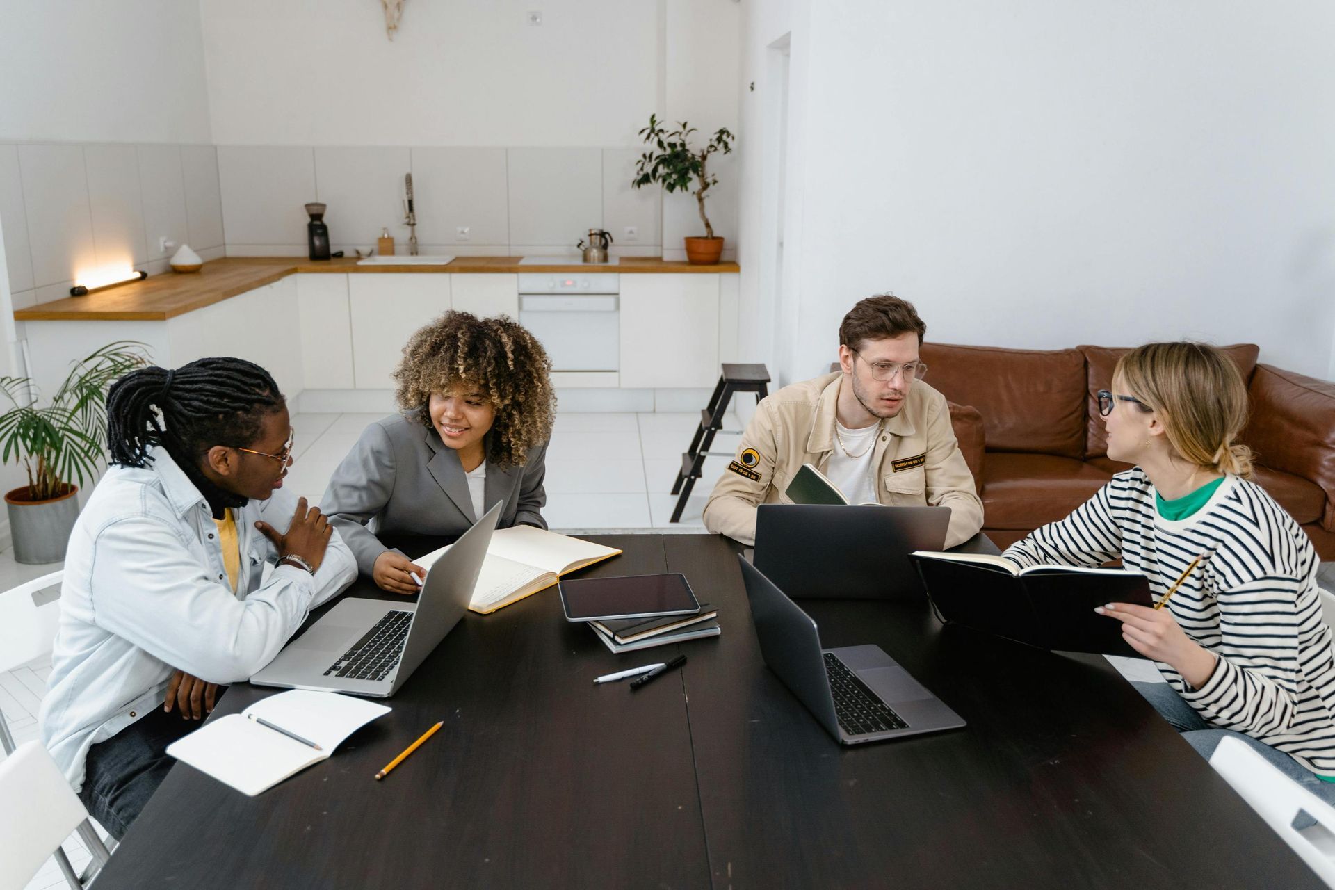 Four young professionals are seated around a large dark table, utilizing laptops and notebooks for a collaborative work or study session. The environment is a modern open-concept room with a white kitchen and a brown leather sofa in the background, blending domestic comfort with a productive workspace.