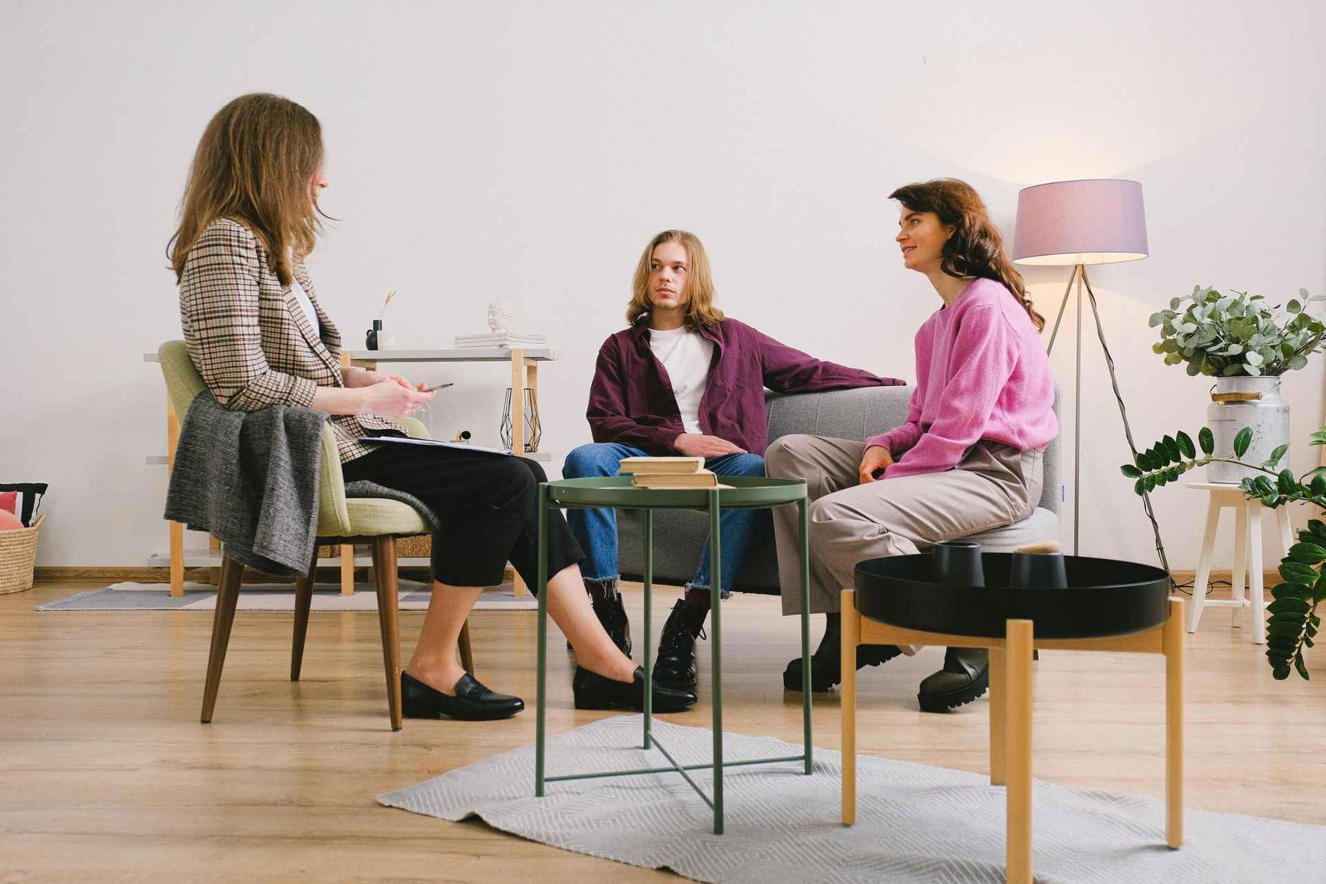 A female counselor sits on a chair facing a young couple seated on a grey sofa, engaging in what appears to be a therapy or mediation session. The minimalist room features simple furniture, a floor lamp, and greenery, providing a calm setting for their focused discussion.