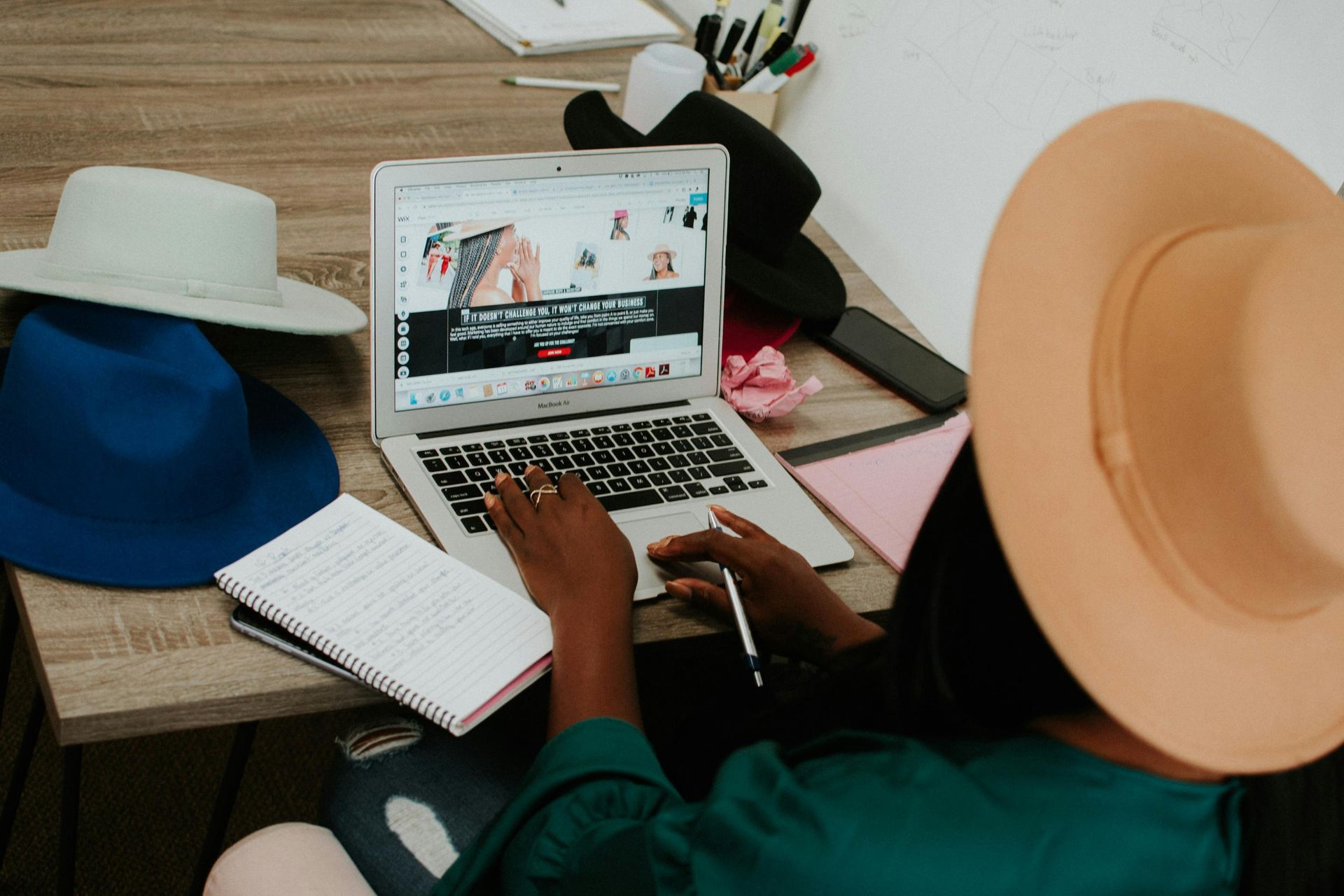 A creative professional, wearing a large tan hat, works diligently on a laptop screen surrounded by several colorful fedoras on her desk. She balances digital work with physical brainstorming, evident from the open spiral notebook and pen in her hand amidst the fashion-focused workspace.