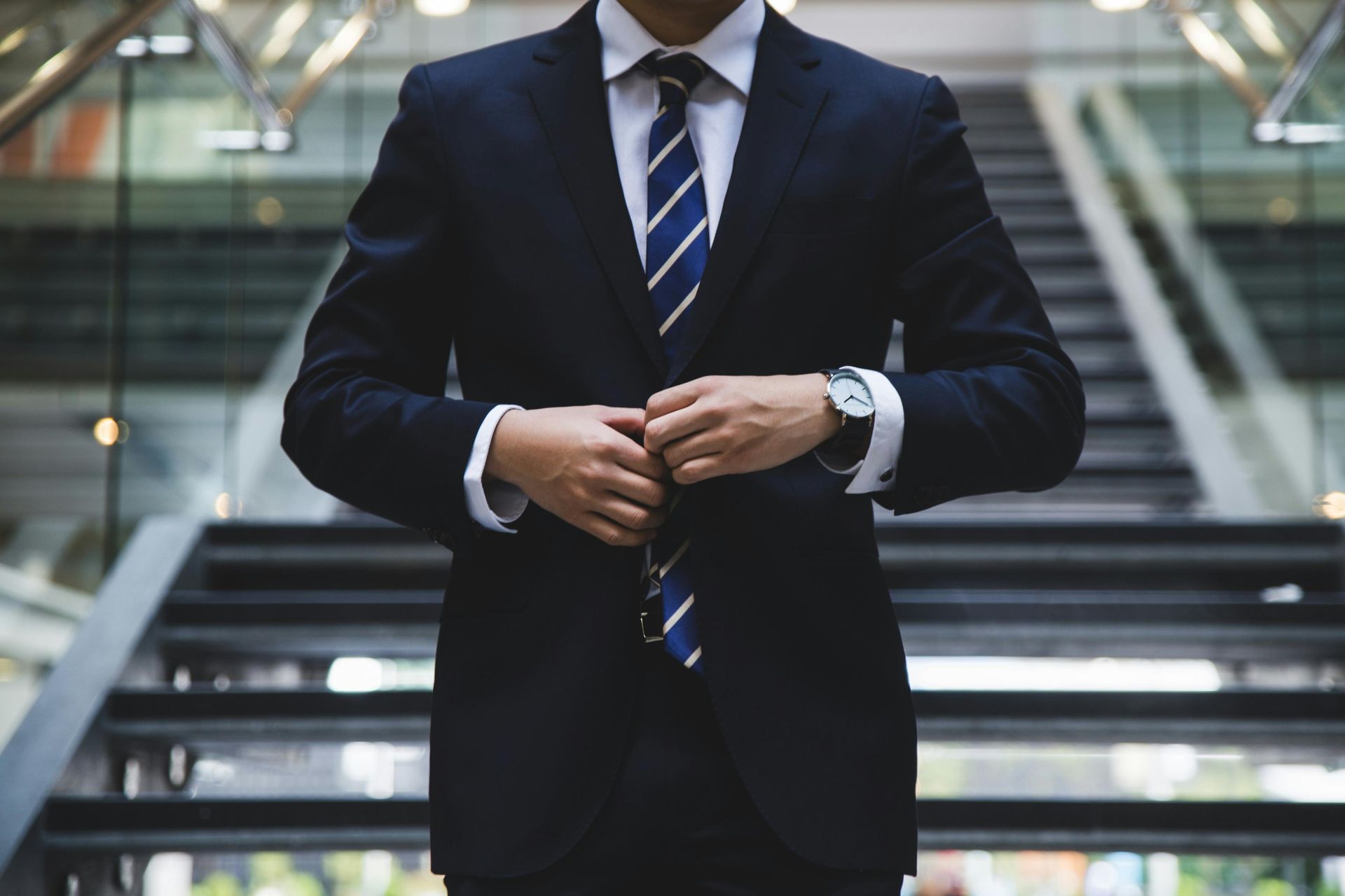 A businessman clad in a dark suit and striped tie stands in front of a modern staircase, adjusting his jacket with both hands. The focus is on his professional attire and posture, suggesting a moment of preparation in a corporate setting.