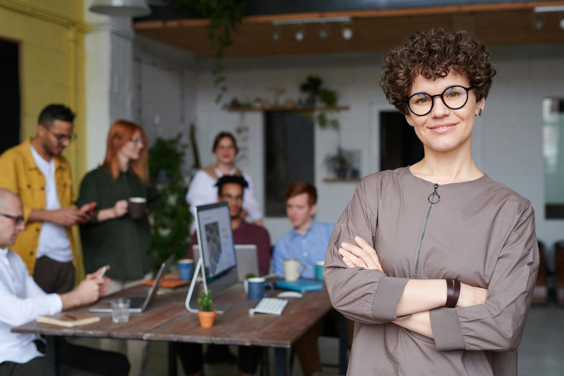 This image features a smiling woman with short curly hair and glasses, standing confidently with her arms crossed in the foreground. Behind her, a busy team of diverse professionals is working together in a casual, open-plan office space.
