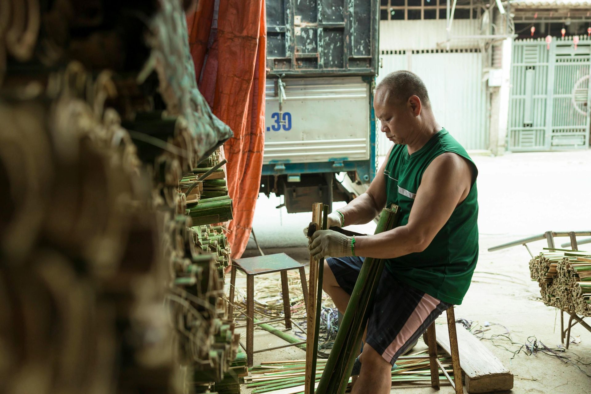 A man wearing a green sleeveless shirt and protective gloves sits on a stool, meticulously splitting long stalks of green bamboo. To his left stands a large stack of processed bamboo segments, while a delivery truck and street gate are visible in the background behind his workspace.