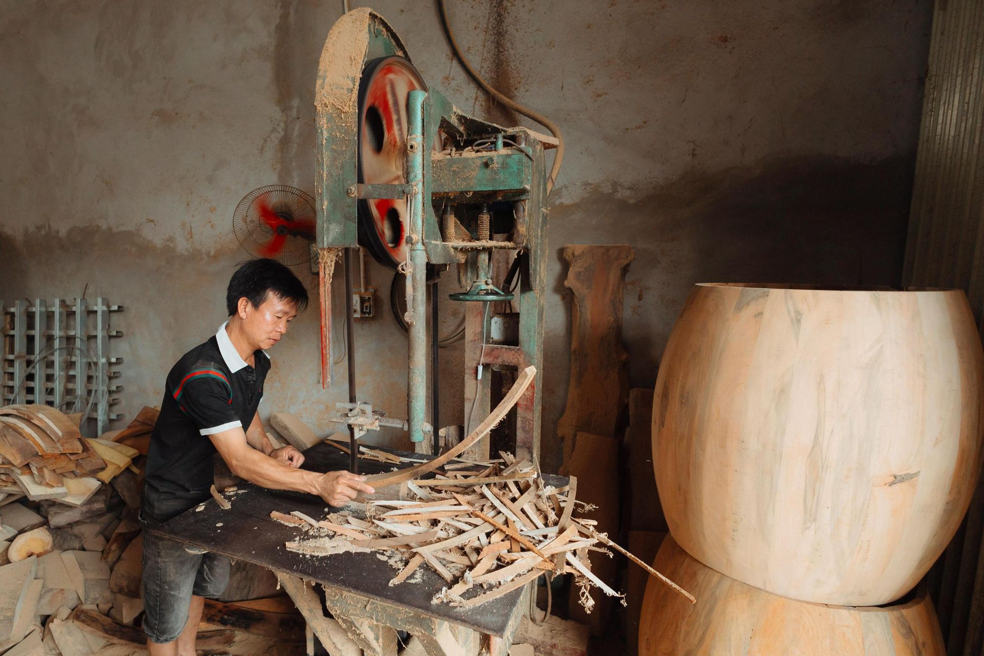 A man stands at a dusty industrial bandsaw in a workshop, carefully guiding a curved piece of wood through the blade. Piles of wood shavings cover his workbench, while large, smooth wooden drum shapes are stacked to his right amidst other lumber scraps.