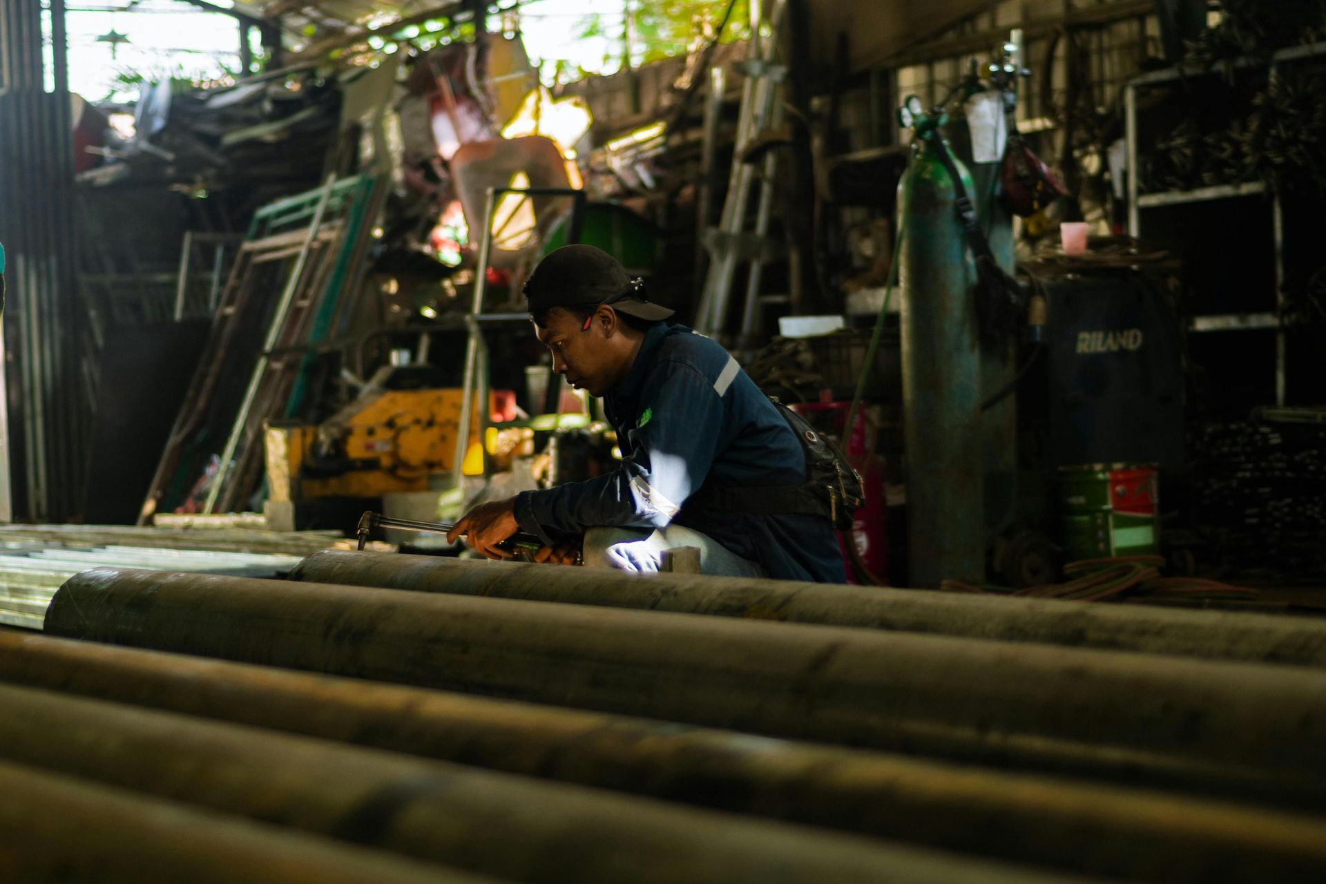 A worker in a blue uniform and cap crouches behind a row of large metal pipes, focusing intently on a task involving a handheld tool. The workshop around him is densely packed with industrial equipment, including metal frames, shelving units, and a large green gas cylinder standing in the dimly lit background.