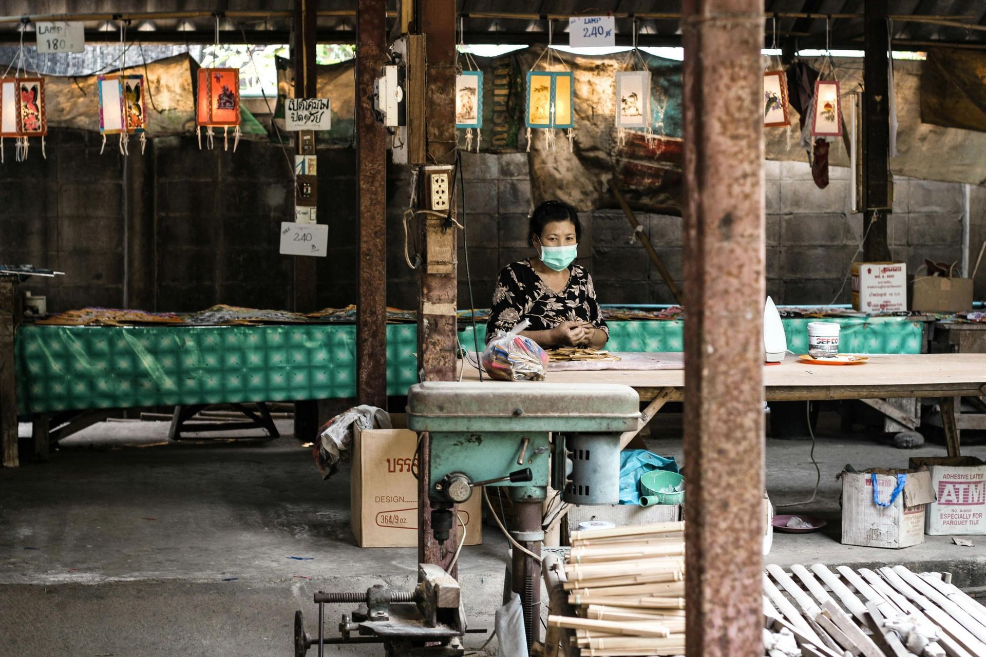 A woman wearing a face mask and floral top sits at a large wooden workbench in an open-air artisanal workshop, pausing to look directly at the camera. The scene is framed by rusty metal pillars and a green drill press in the foreground, while a row of decorative paper lanterns hangs in the background behind her.