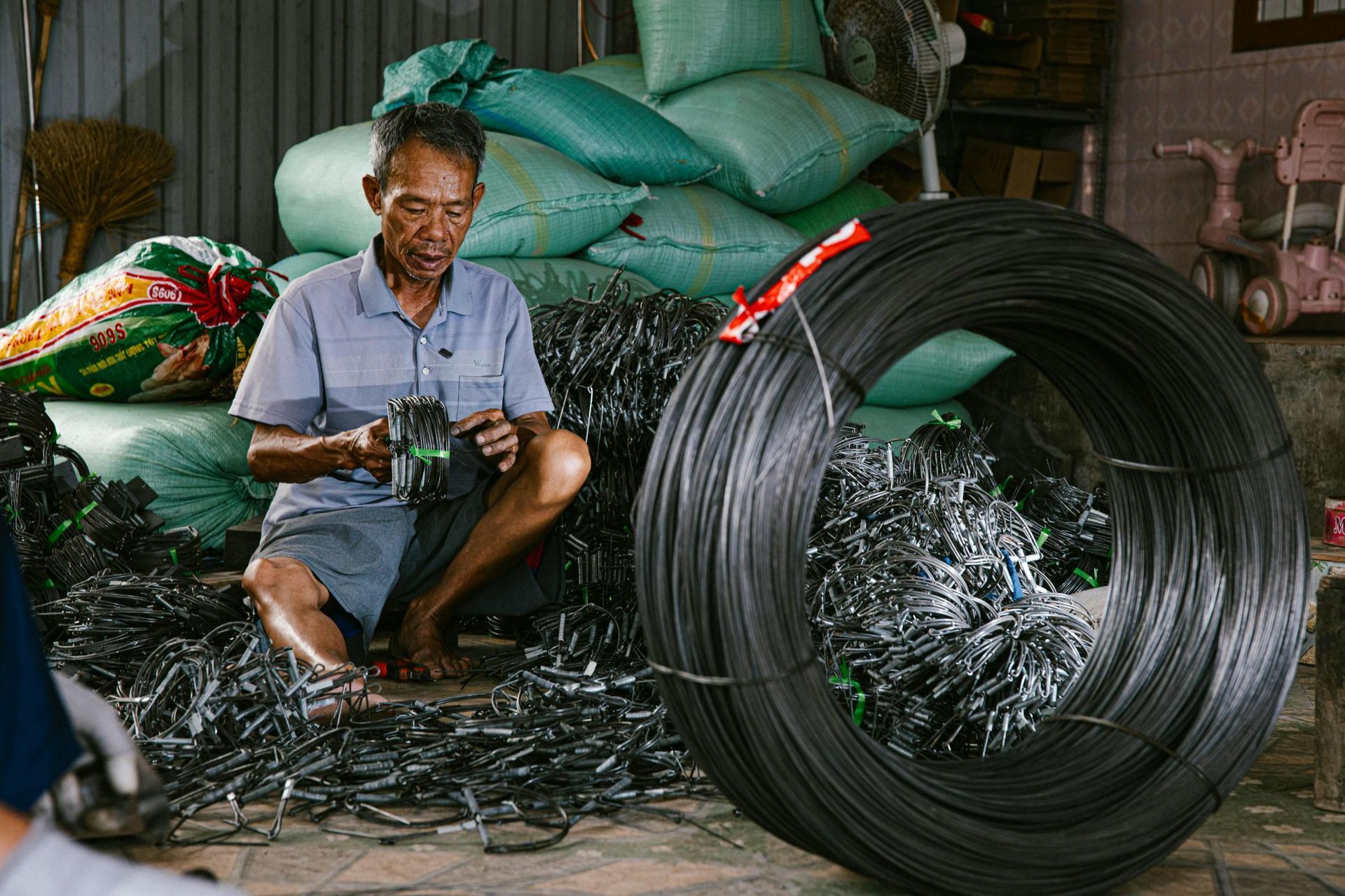 An older man sits barefoot on the floor of a workshop, diligently bundling small metal wire loops by