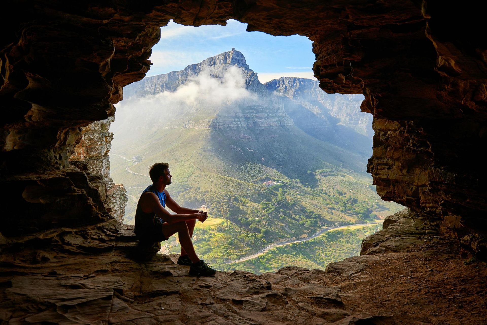 A man sits at the edge of a rocky cave opening, looking out over a vast landscape dominated by a maj