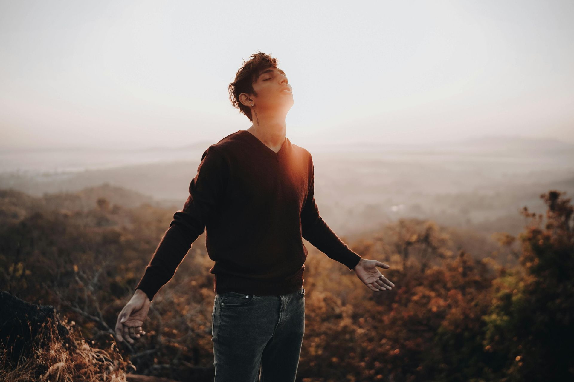 A young man stands with his head tilted back and arms slightly outstretched, basking in the warm, golden light of the sun. Behind him, a hazy landscape of rolling hills and trees stretches out, creating a serene and peaceful atmosphere.