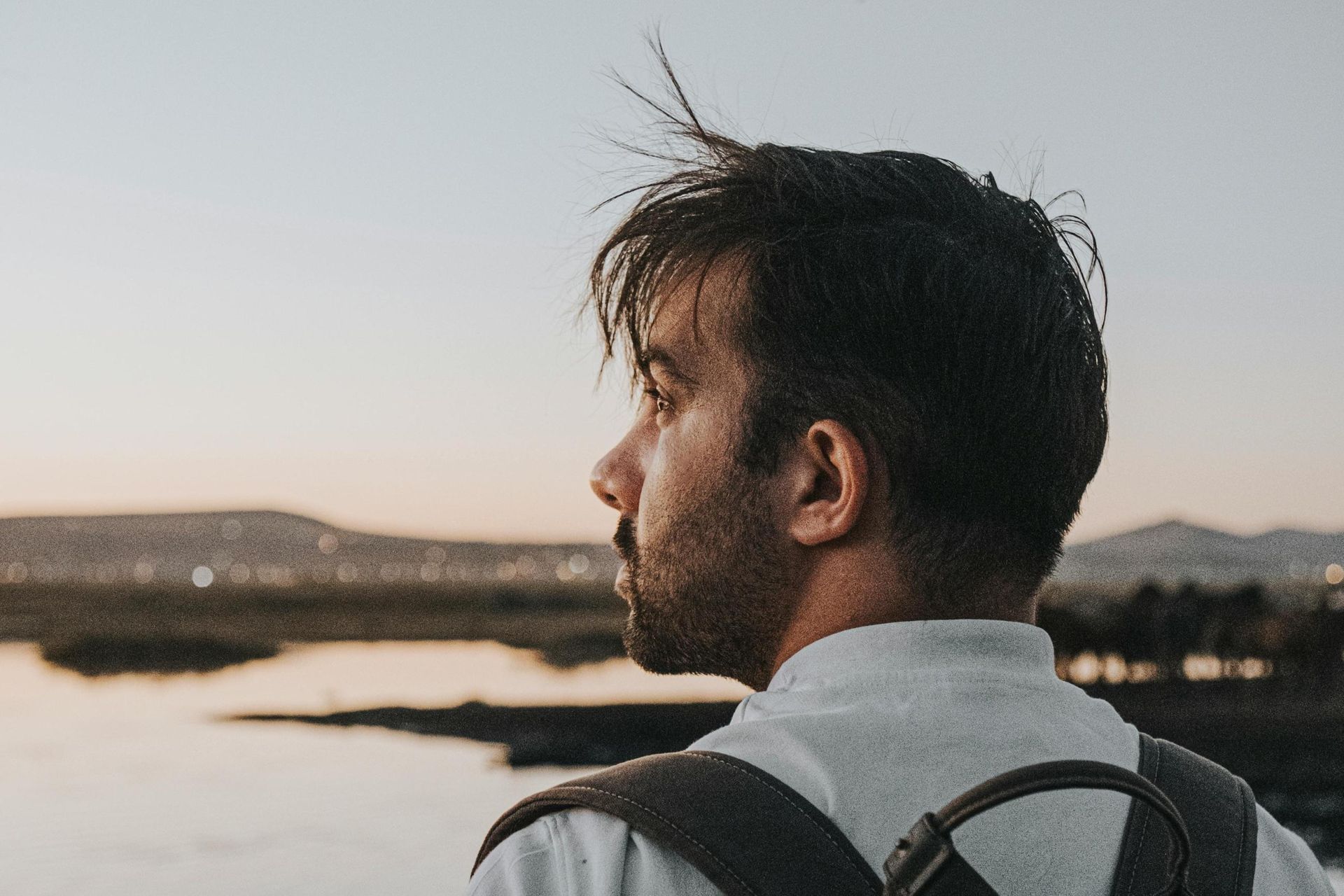 A man wearing a backpack stands in profile, looking out over a calm body of water towards distant hi