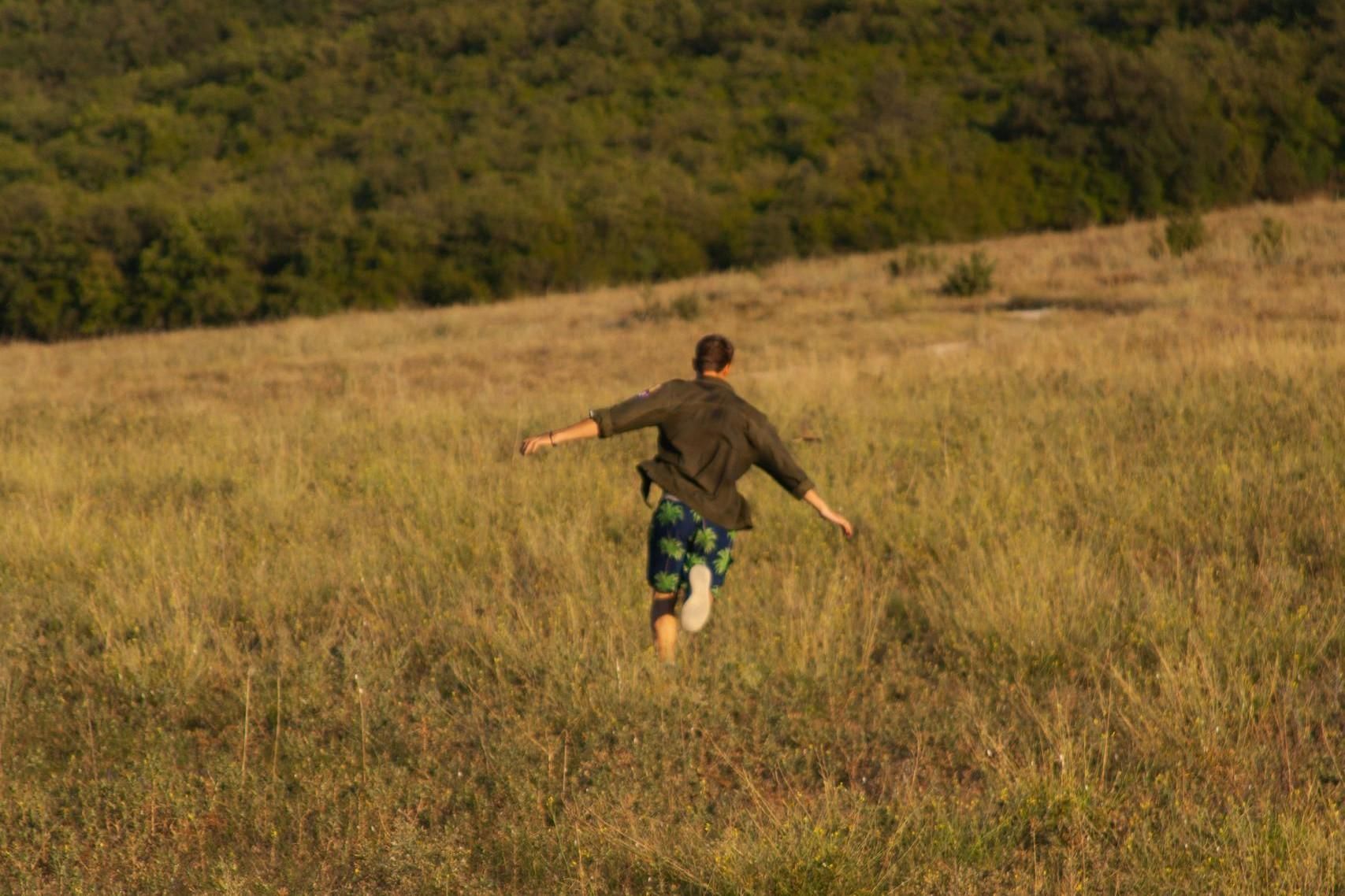 A man runs away from the viewer through a field of tall, golden grass with his arms widely outstretched. Wearing patterned blue shorts and a dark shirt, he heads towards a dense, green forest that rises in the background.