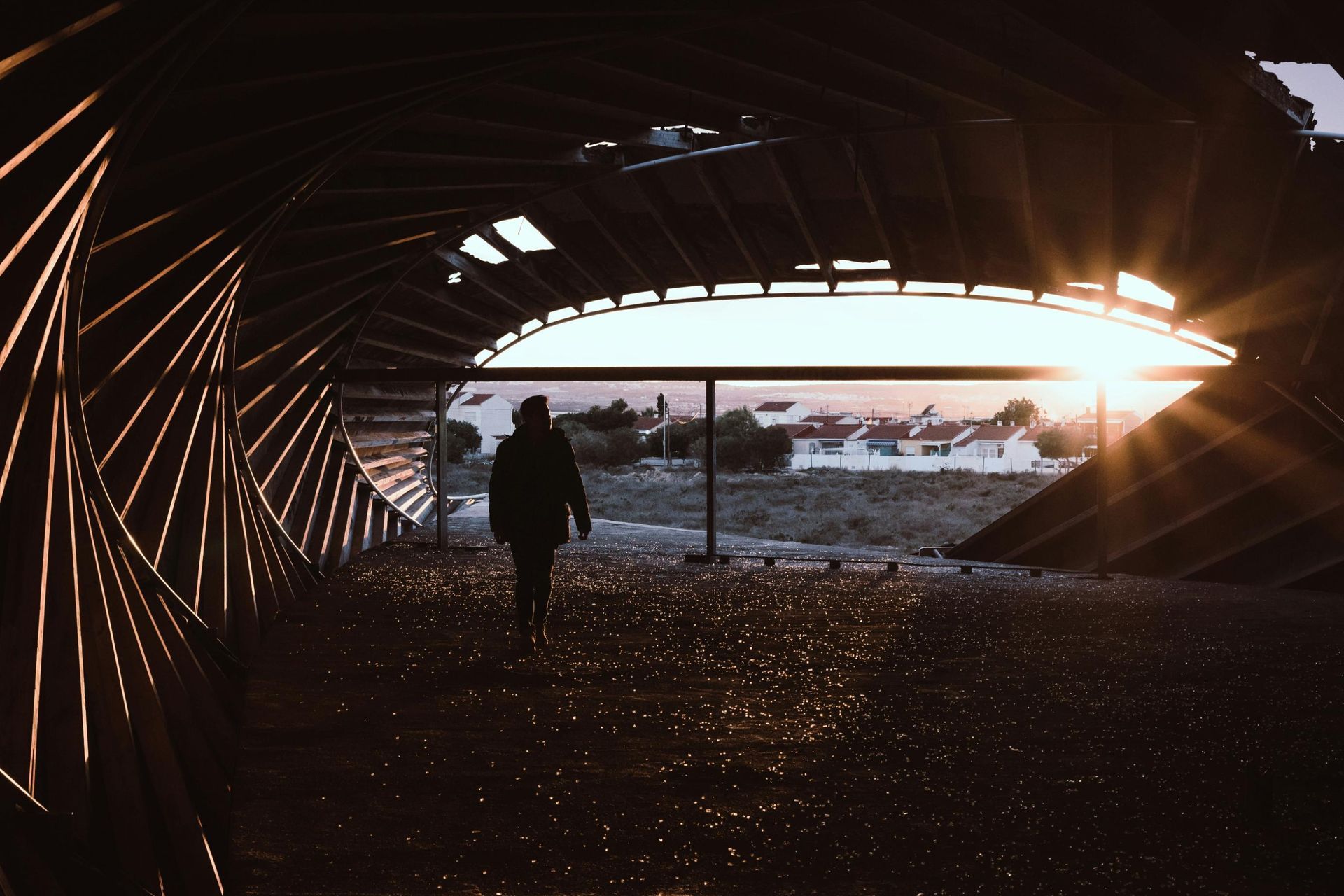 A silhouetted figure walks through a cavernous, spiraling structure made of dark, angled beams. Sunlight bursts through the wide opening at the end, casting golden sparkles on the ground and framing a distant townscape.