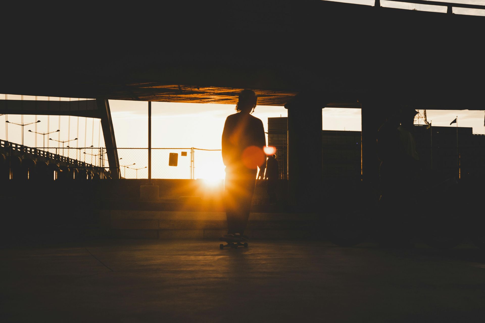 A silhouetted figure stands on a skateboard beneath the dark, concrete span of a bridge or overpass. The bright sun bursts through the background, creating a dramatic flare and casting a warm, golden glow across the urban scene.