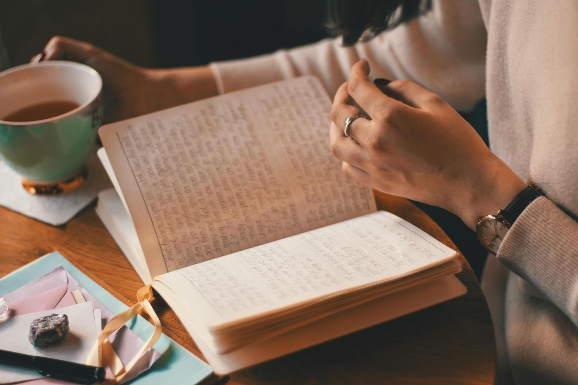 A person wearing a watch and ring sits at a wooden table, deeply engaged in reading an open journal filled with handwritten entries. Beside the notebook sits a light green teacup, creating a peaceful and reflective atmosphere for journaling or study.