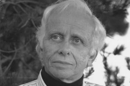 This black-and-white portrait depicts George Leonard with white hair, gazing intently off to the side with a serious expression. He is dressed in a striped button-down shirt layered under a dark sweater, standing against a blurred backdrop of pine branches.