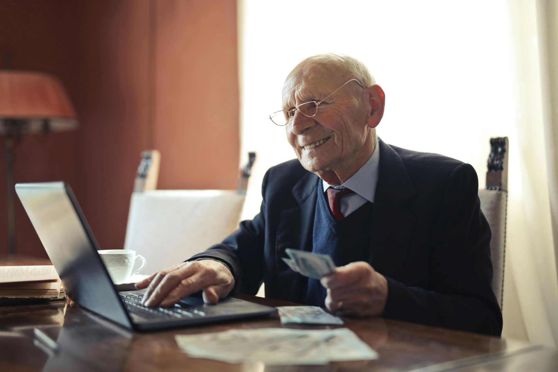 A smiling elderly man dressed in a formal suit sits at a table, holding a stack of cash while active