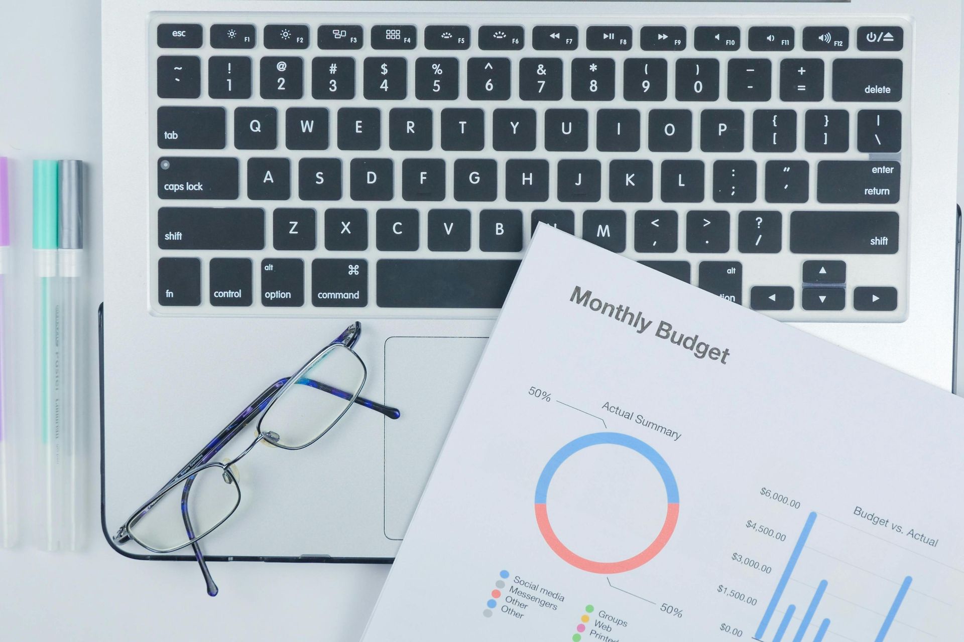 A top-down shot features a laptop keyboard with a pair of reading glasses resting on the trackpad and a printed document titled 
