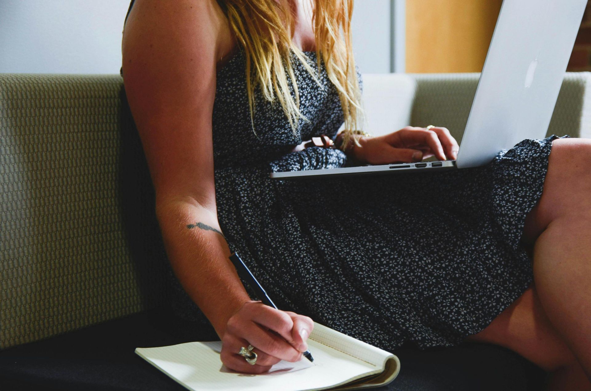 A young woman sits on a sofa, balancing a laptop on her legs while simultaneously writing notes in a physical notebook. Dressed in a sleeveless floral dress, she appears to be multitasking in a relaxed, casual environment.