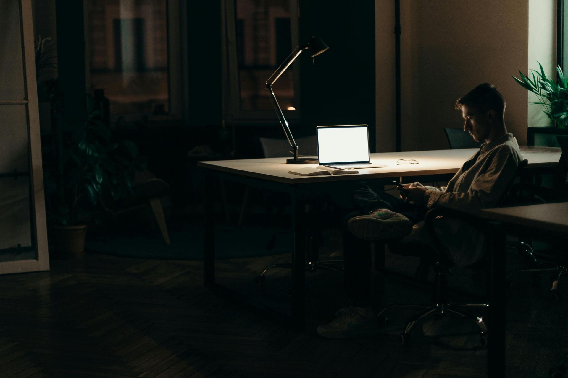 A solitary man sits at a desk in a dimly lit office, illuminated chiefly by a desk lamp and the glow