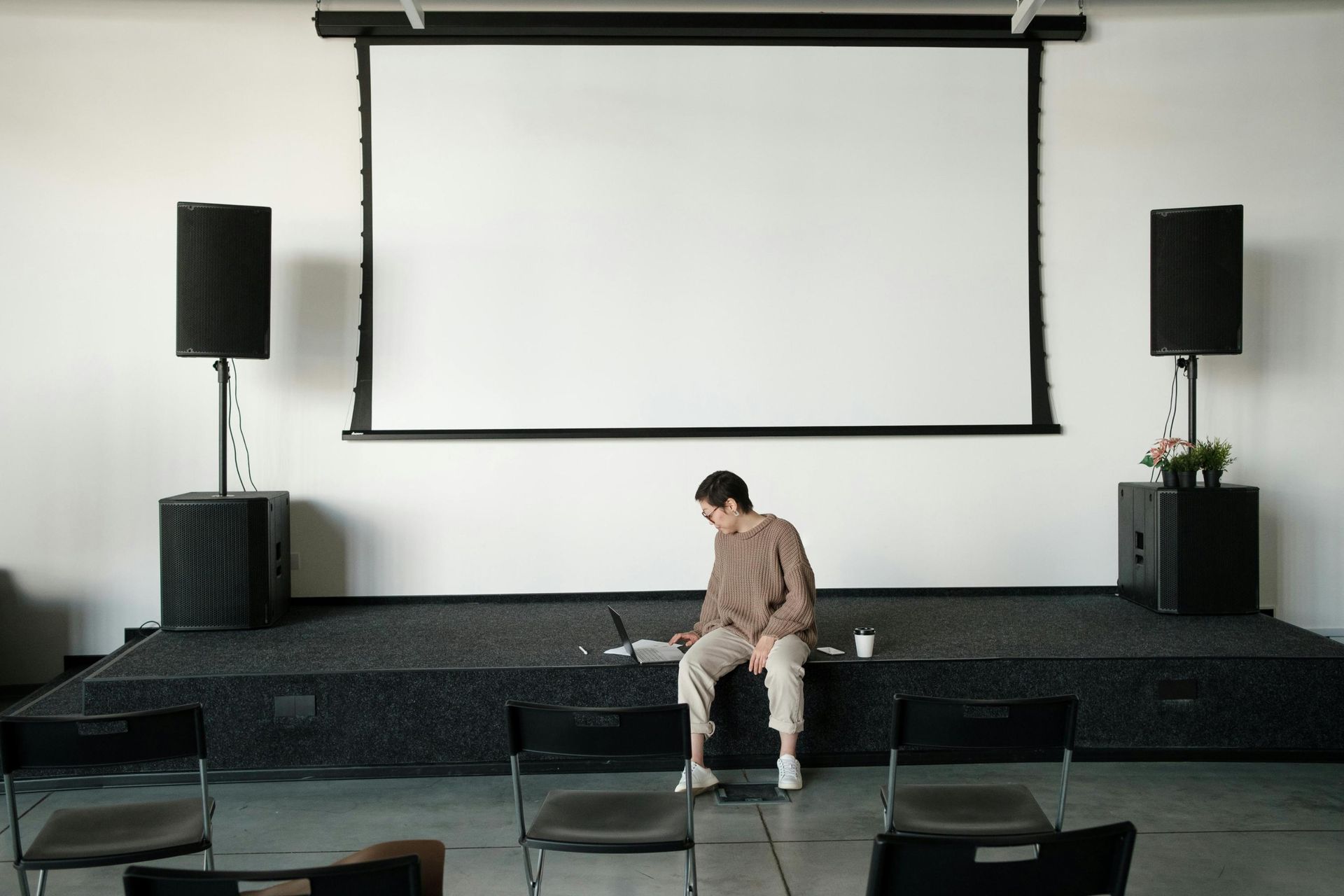 A person sits quietly on the edge of a stage platform, working on a laptop beneath a large, blank projection screen. Flanked by tall black speakers and facing rows of empty chairs, the scene captures a moment of solitary preparation before an event or presentation begins.