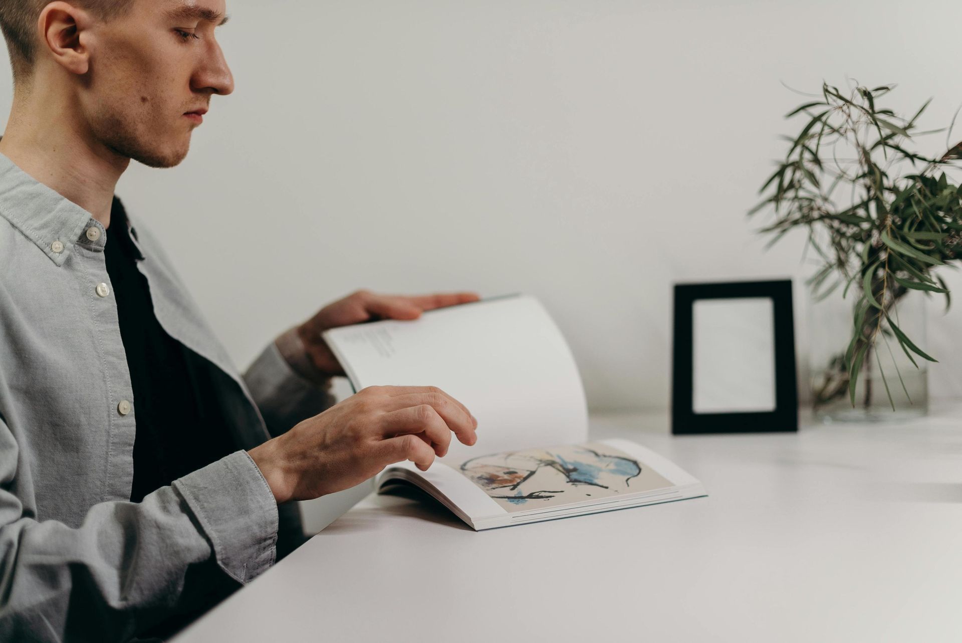 A focused young man sits at a clean white desk, turning the pages of an open book that features colorful illustrations. To his side, a minimalist arrangement includes a black picture frame and a glass vase filled with greenery.