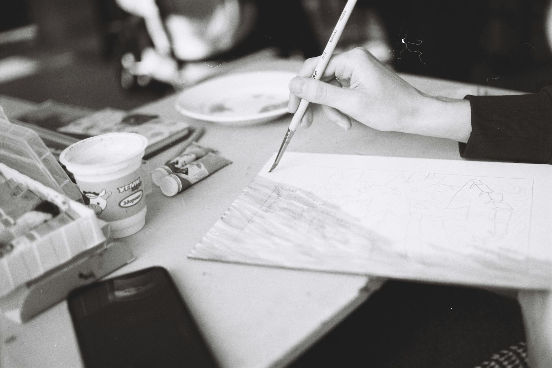 This black-and-white photograph captures an artist's hand in the process of painting over a pencil sketch on a sheet of paper. The table is cluttered with creative supplies, including small tubes of paint, a water cup, and a smartphone lying nearby.