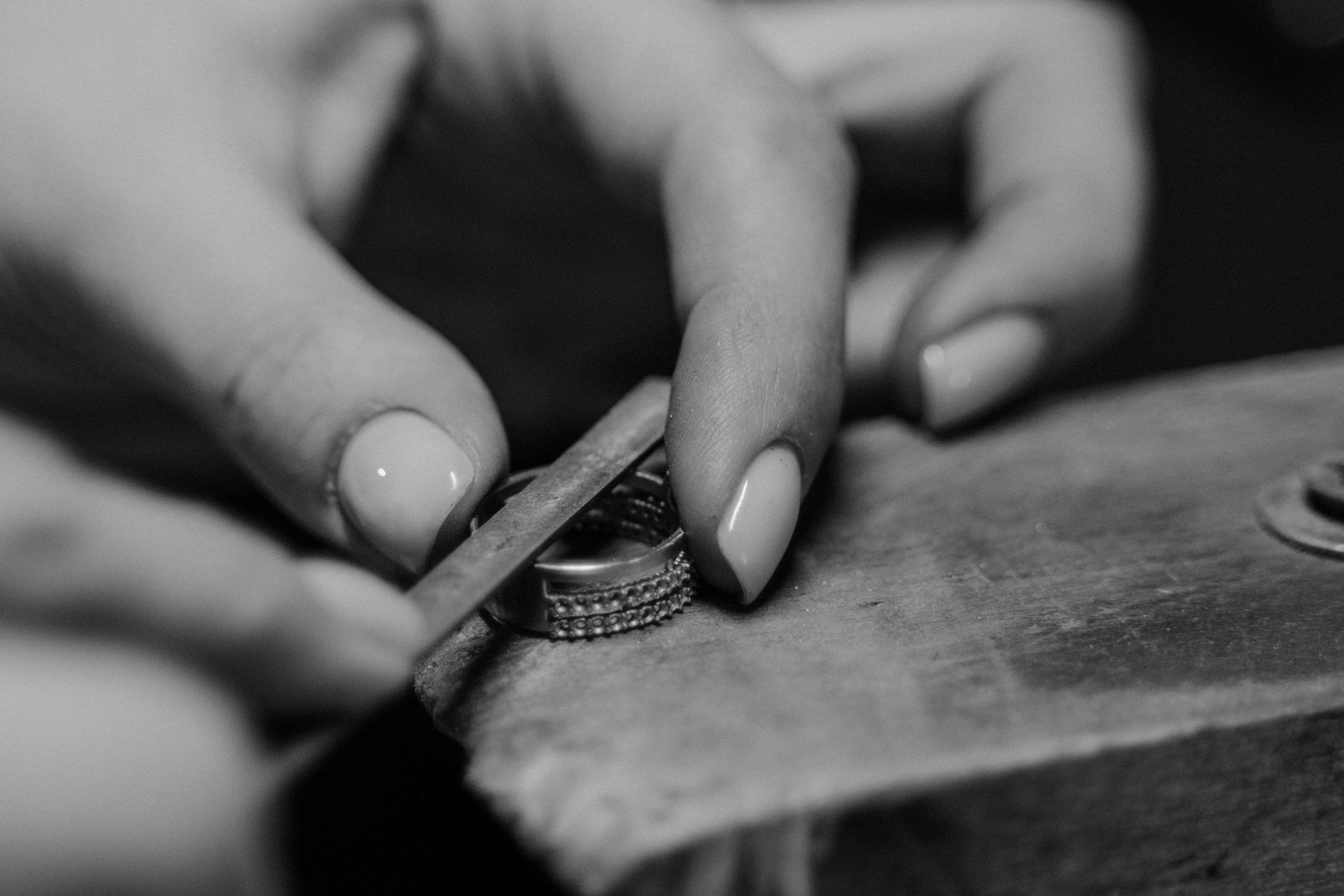 In this close-up black-and-white photograph, a pair of hands is shown delicately working on a studded ring using a flat metal tool. Resting on a worn wooden block, the hands demonstrate precise craftsmanship as they shape or finish the jewelry piece.