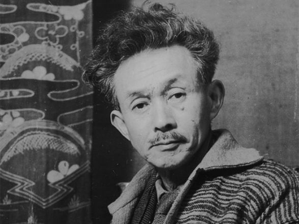 This black-and-white portrait features Soetsu Yanagi, the author of the previously mentioned book, gazing directly at the viewer with a calm, contemplative expression. He is wearing a textured, patterned jacket and is seated in front of a hanging fabric adorned with traditional Japanese motifs, including stylized pine trees and floral patterns.