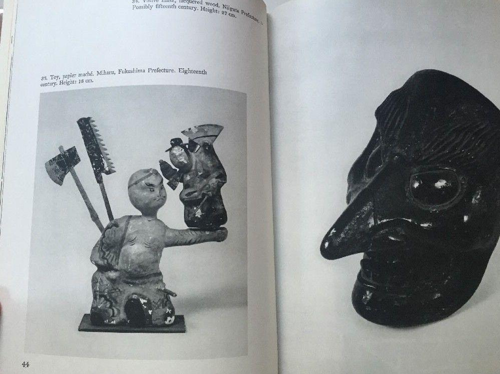 This image displays an open spread of the book featuring black-and-white photographs of traditional artifacts, with the left page showcasing an 18th-century papier-mâché toy from Fukushima Prefecture. The right page presents a striking close-up of a dark, glossy mask with an elongated nose, contrasting with the smaller figure opposite it.