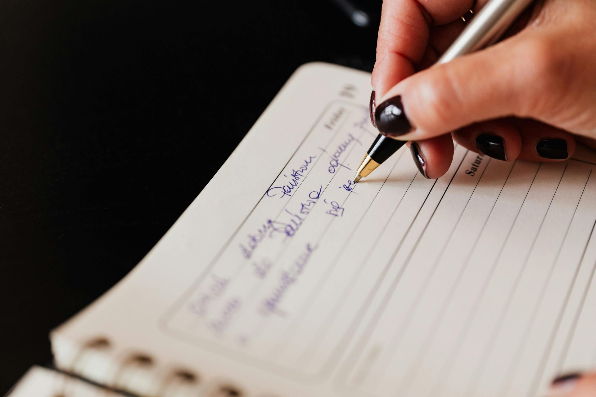 A person with dark-polished fingernails holds a silver and black pen while writing in a spiral-bound notebook. The handwriting is in blue ink on lined paper, which appears to be part of a daily planner or journal.