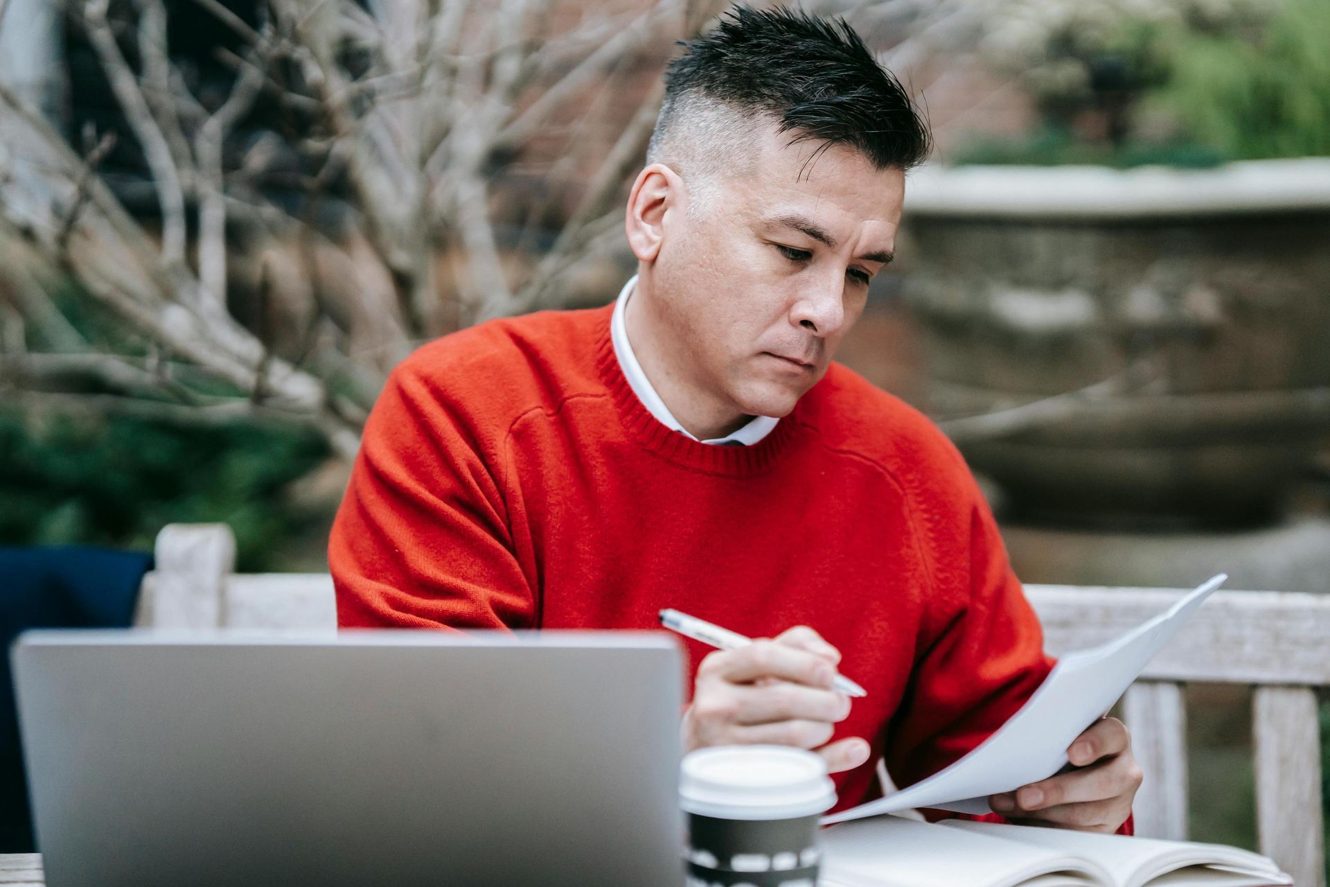 A man in a vibrant red sweater sits at an outdoor table, focused on reviewing documents while holding a pen. A laptop, an open book, and a coffee cup are spread out before him, suggesting a productive work session in a garden-like setting.