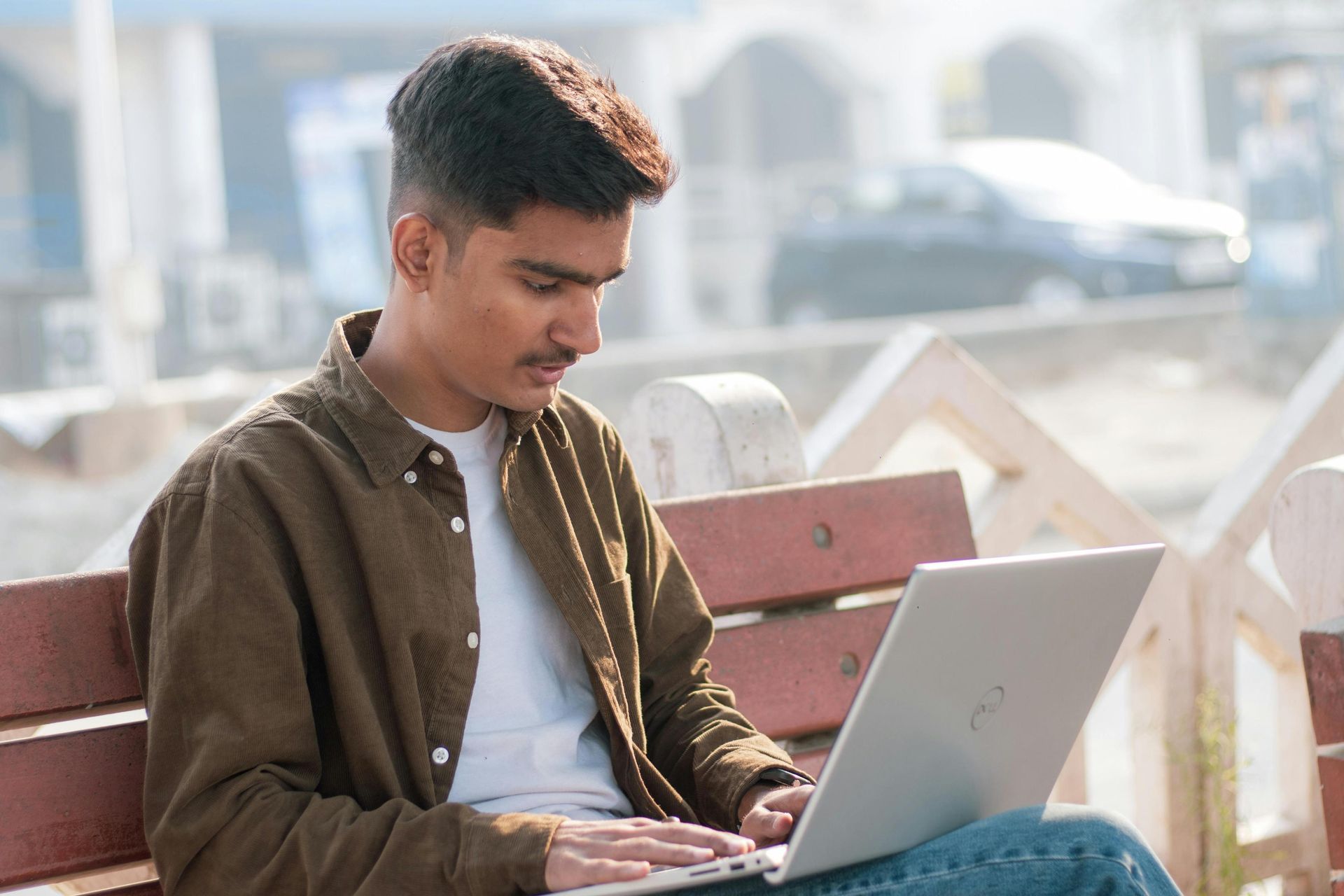 A young man wearing a brown corduroy shirt over a white t-shirt sits on an outdoor bench while focused on his laptop. The background shows a bright, slightly blurred urban setting with the suggestion of a street and parked vehicles.