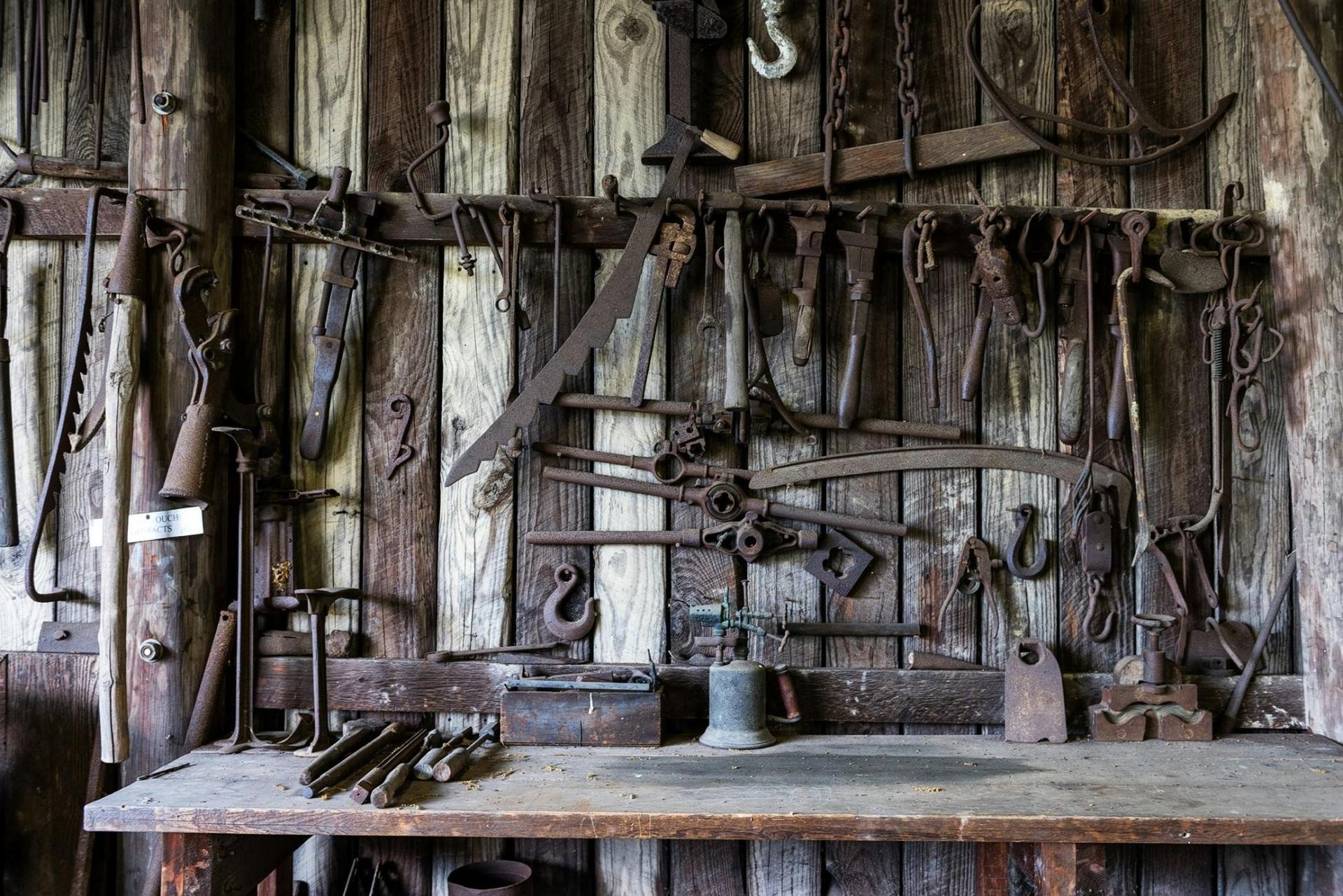 A vast collection of antique, rusted tools hangs organized against a weathered wooden wall, evoking the atmosphere of a historic workshop or barn. Below the hanging display, a sturdy wooden workbench holds additional implements like chisels and a vintage torch, highlighting a scene rich in texture and utilitarian history.