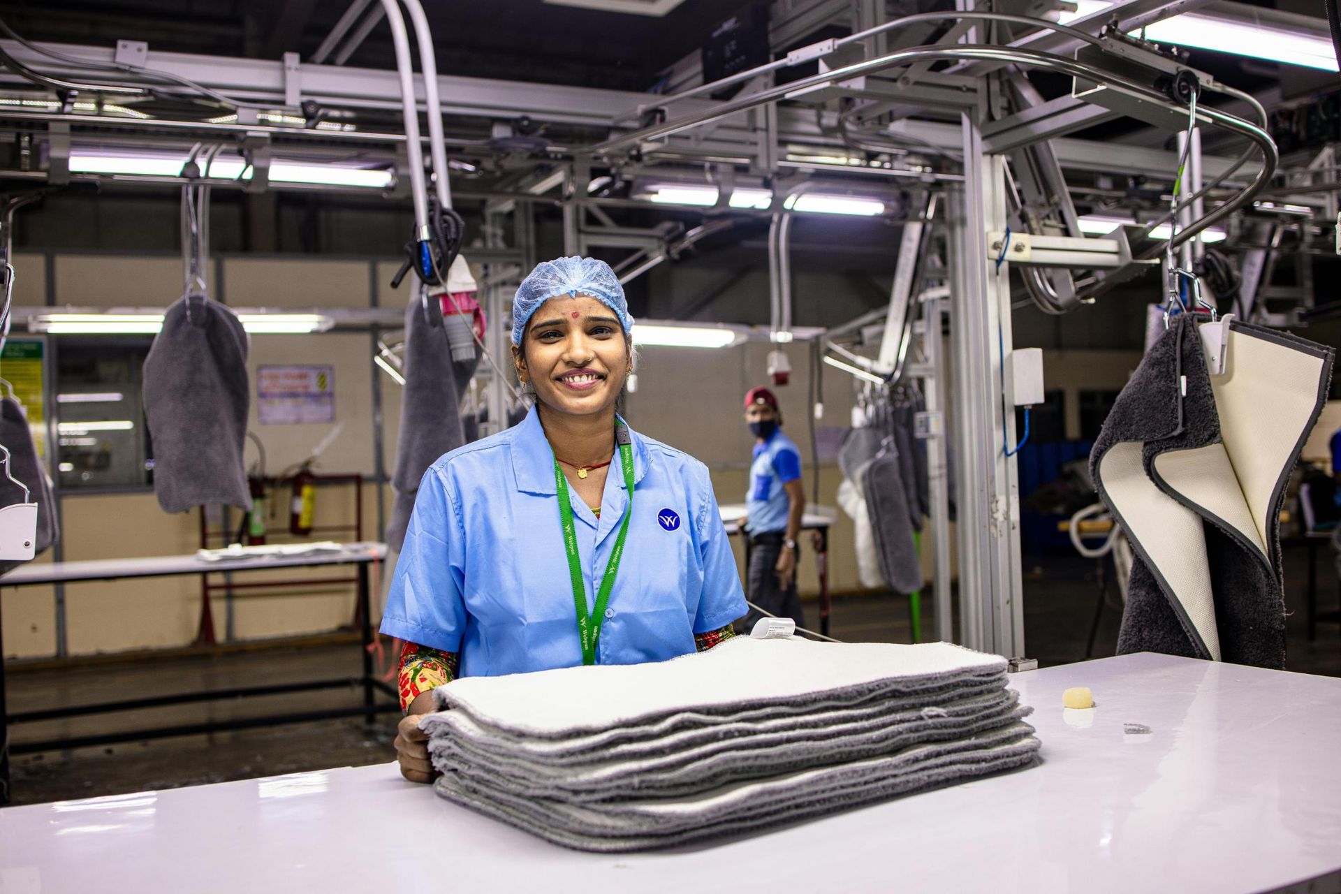 A smiling factory worker in a blue uniform and protective hairnet stands at a table, posing behind a neat stack of gray and white textile materials. Behind her, the industrial environment features overhead conveyor systems and bright lighting, highlighting a busy manufacturing setting with another employee working in the background.