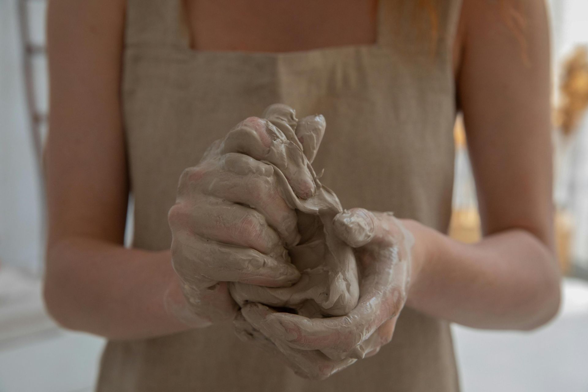 In a close-up shot, a person's hands are deeply engaged in kneading and shaping a malleable lump of wet clay. The artisan wears a beige apron that blends with the earthy tones of the scene, highlighting the tactile and messy process of pottery creation.