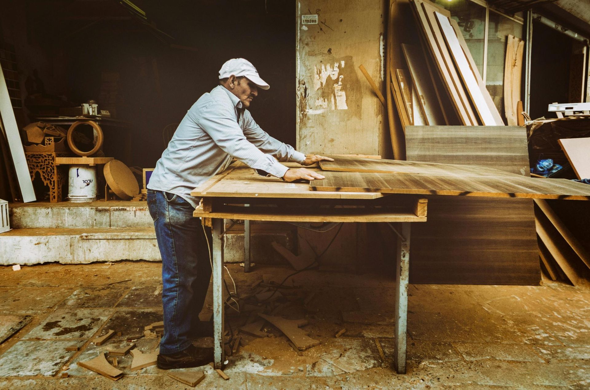 A carpenter wearing a white cap and button-down shirt stands at a work table, carefully positioning a large sheet of wood. The surrounding workshop is filled with stacks of lumber and scattered wood scraps on the dusty floor, suggesting a busy and productive environment.