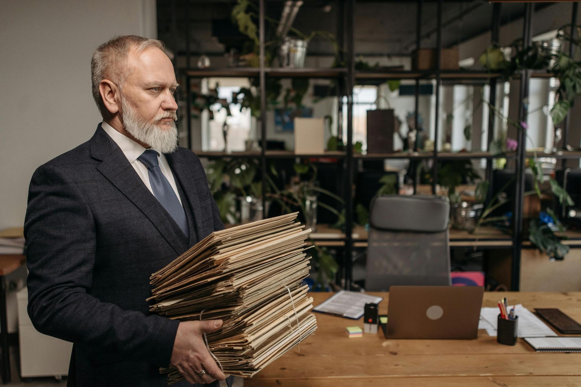 A distinguished older man with a grey beard and dark suit stands holding a massive, heavy stack of paper files. The setting appears to be a modern office, with a wooden desk, laptop, and plant-filled shelving unit visible in the background.