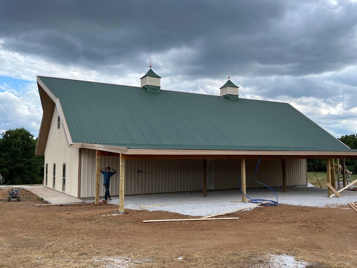 A large barn with a green roof is being built in a dirt field.