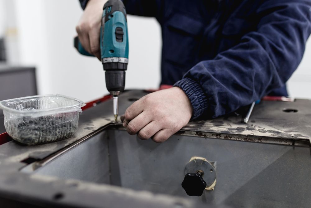 Una persona usa un taladro eléctrico para fijar una pieza a una superficie metálica. Cerca hay un contenedor de tornillos.