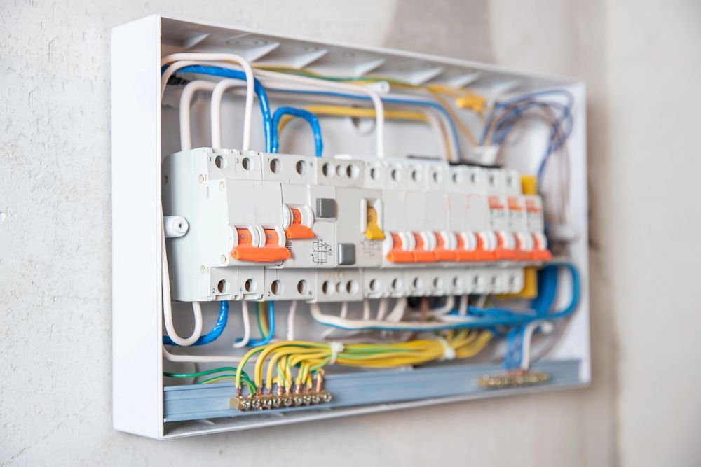 Panel eléctrico abierto con cables, disyuntores y terminales de colores, montado en una pared de color claro.