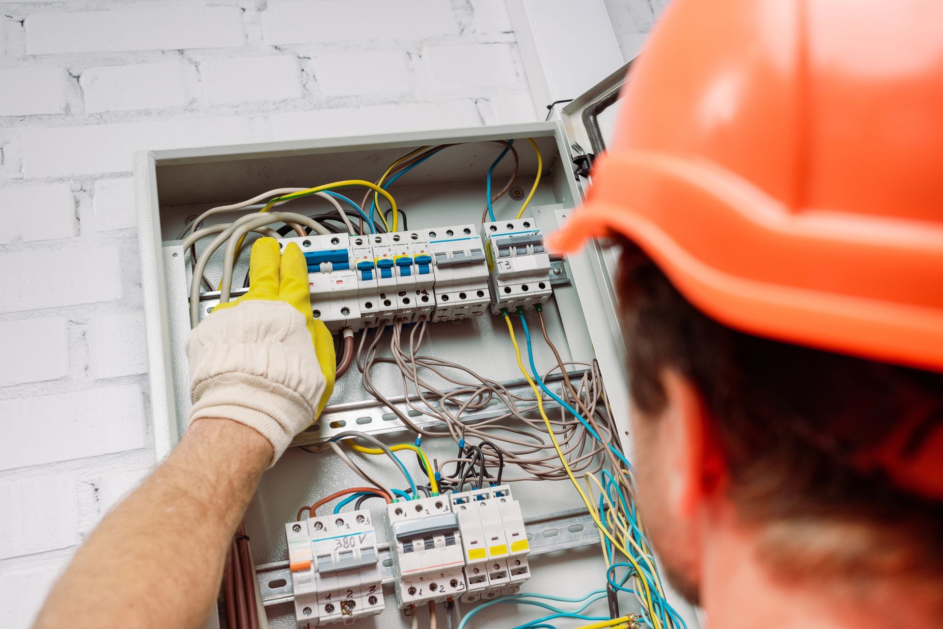 Electricista con casco y guantes de color naranja trabajando en un panel de disyuntores.