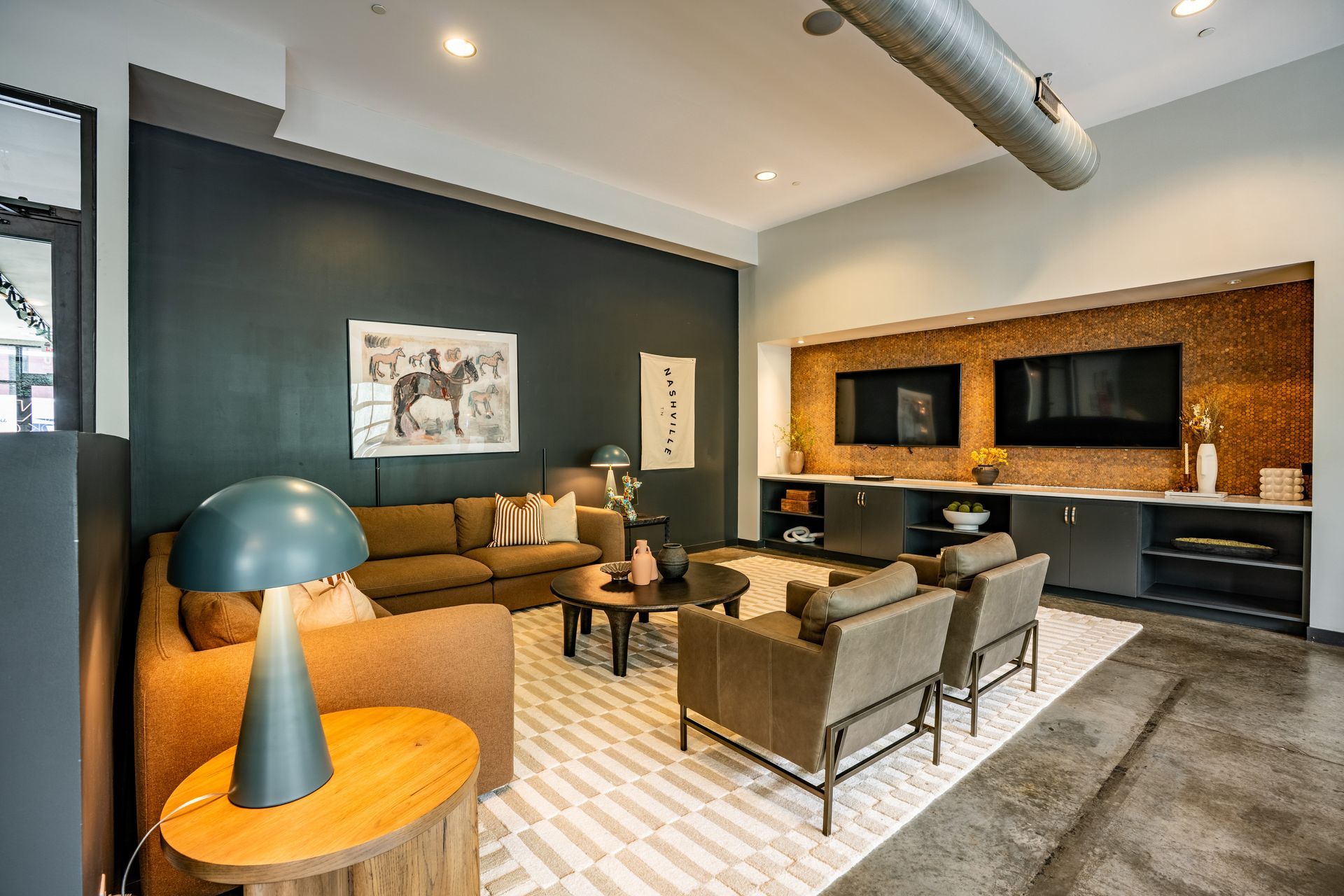 A modern living room with brown furniture, a blue lamp, and two TVs on a textured wall, with a concrete floor.