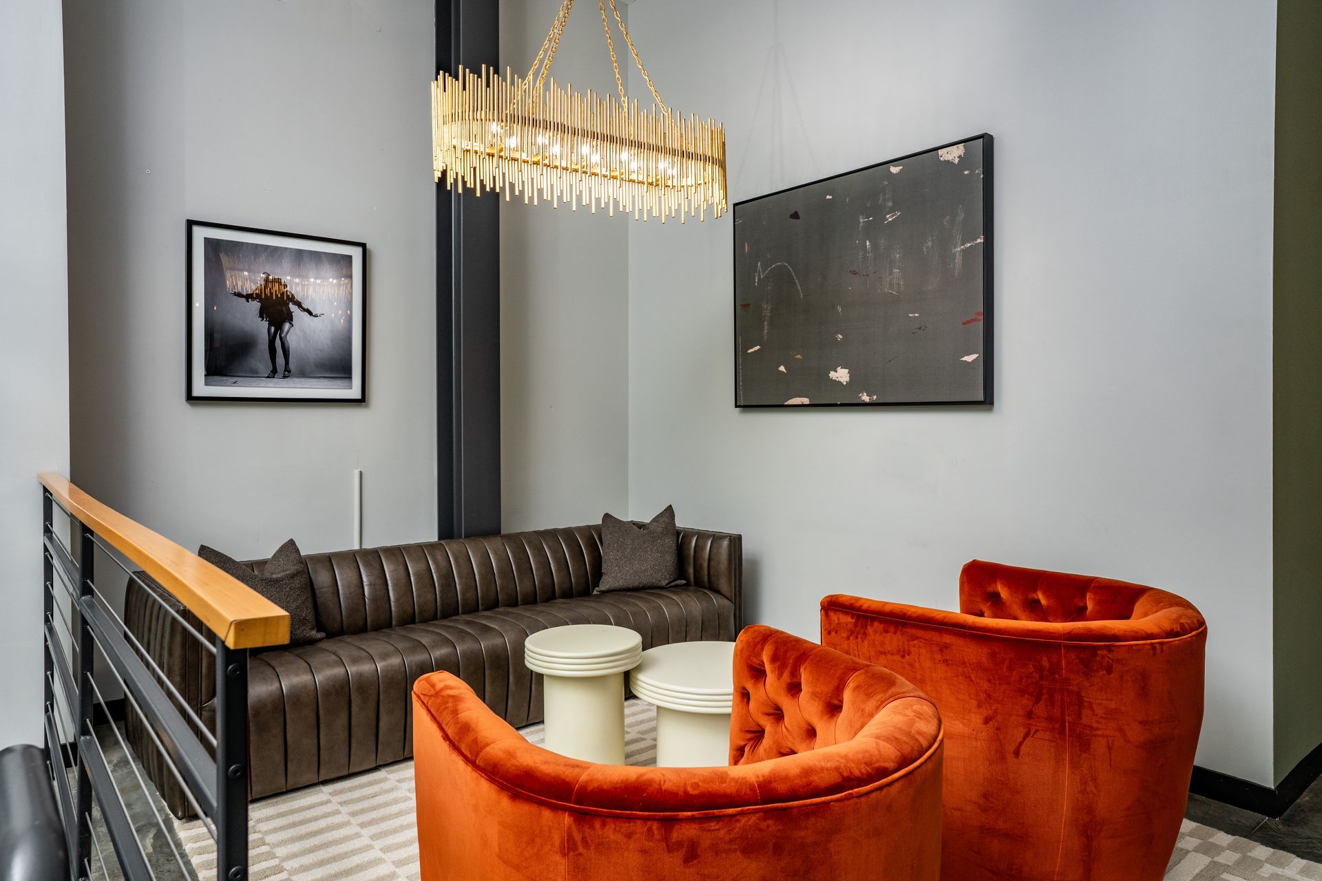 Cozy seating area with a brown leather sofa, two orange armchairs, and artwork on gray walls. Gold chandelier hangs above.