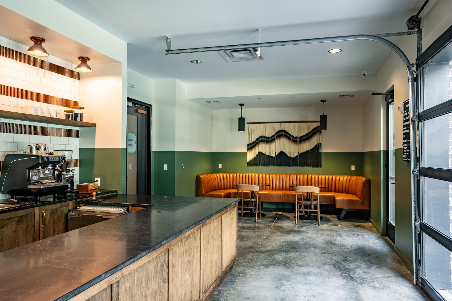 Coffee shop interior: bar, seating area with orange sofa, and a partially open garage door. Green and white walls.